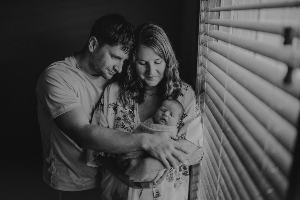 Intimate black and white family portrait near window showing parents tenderly holding their newborn baby.