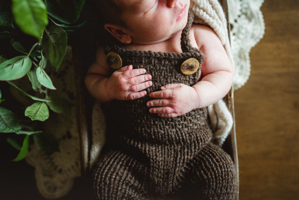 Close-up of infant wearing brown knitted romper with decorative buttons near green plants.
