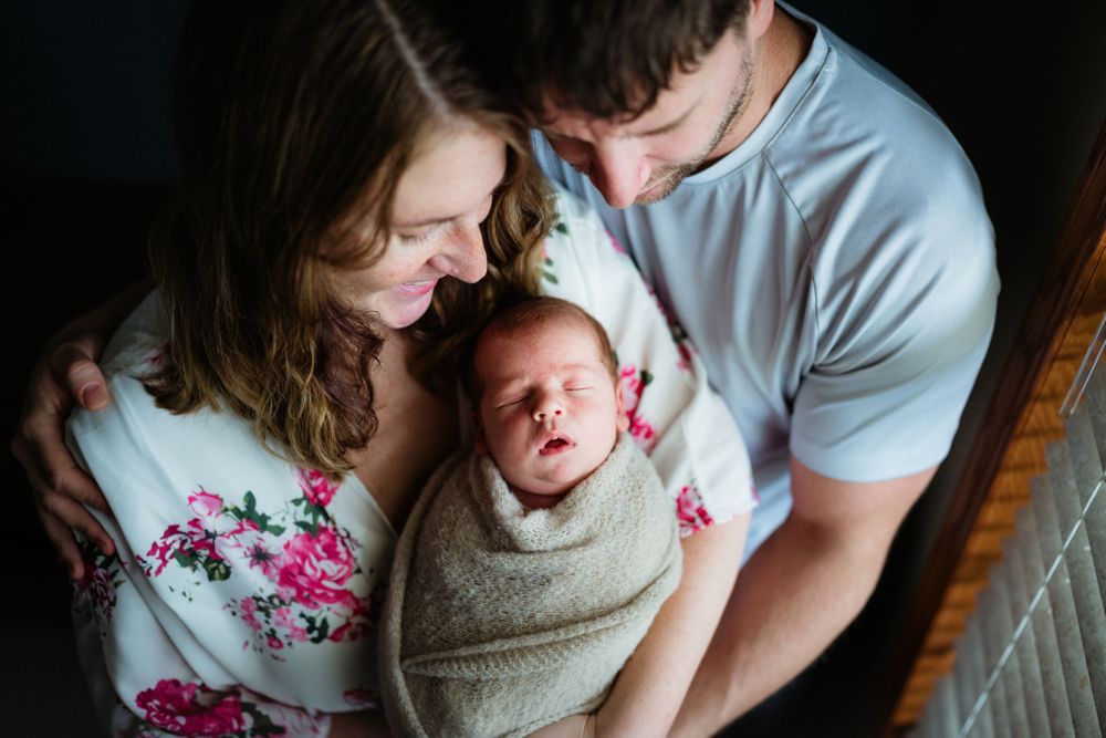 Intimate family portrait with parents cradling swaddled newborn by window.