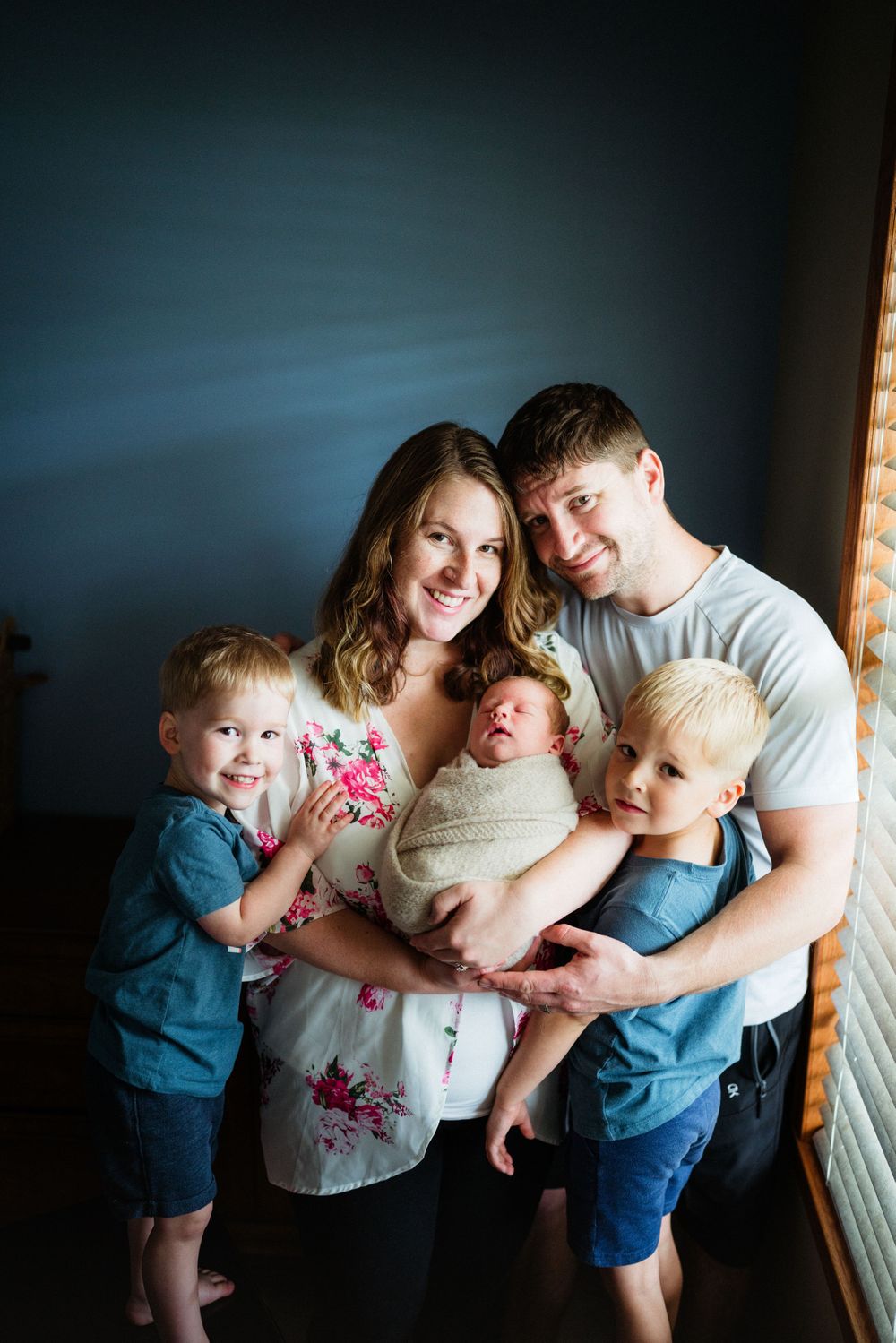 Family portrait showing parents with three children against blue wall and natural window light.