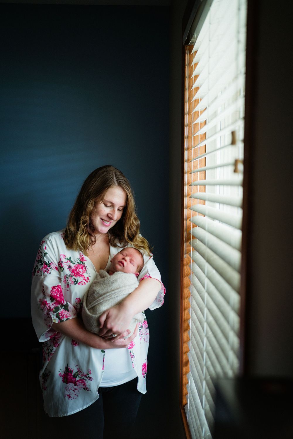 Mother in floral robe holding swaddled newborn by window with streaming light.