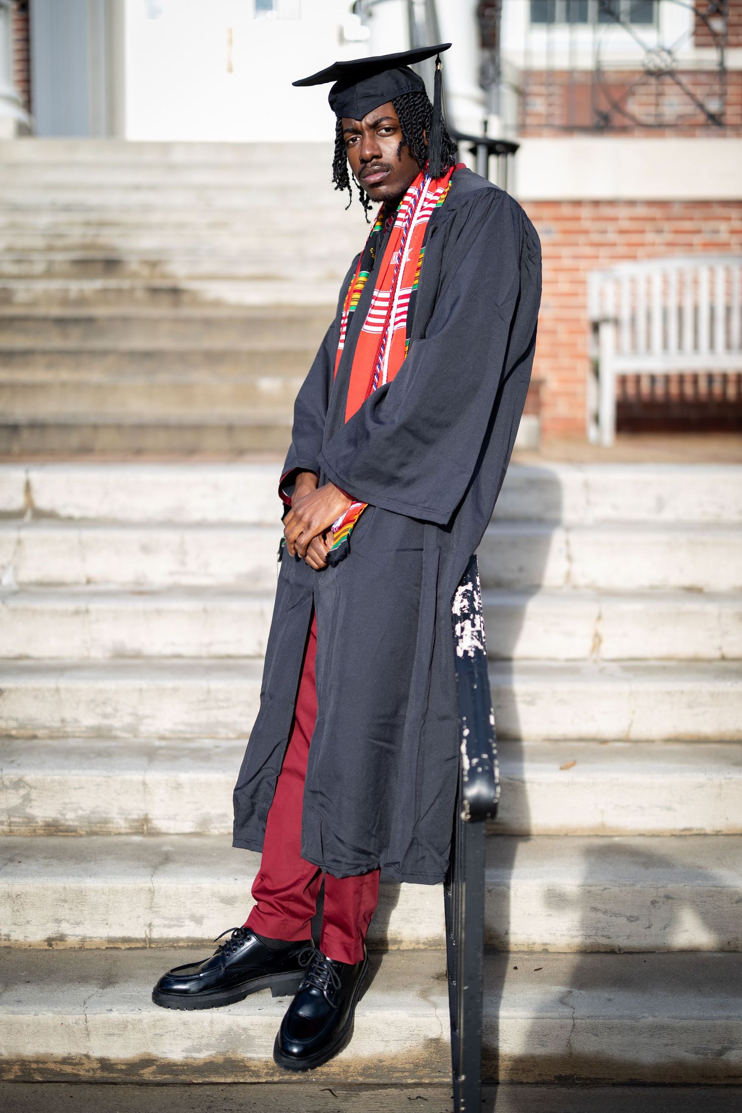 Graduate in traditional black cap and gown with red stole standing on stone steps.