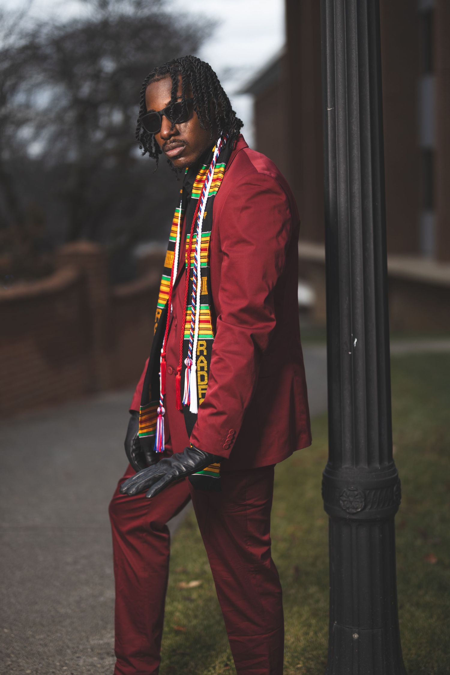 Stylish graduate in burgundy suit and patterned scarf posing by lamp post.