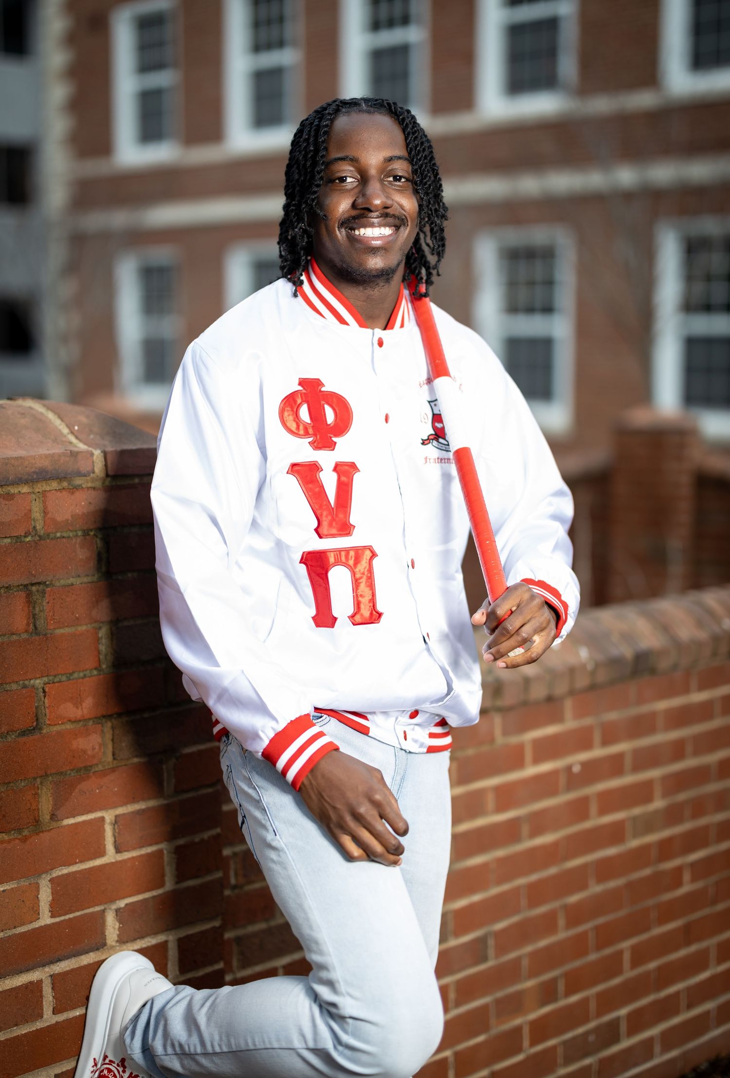Graduate in white outfit with red accents leaning against brick wall outdoors.