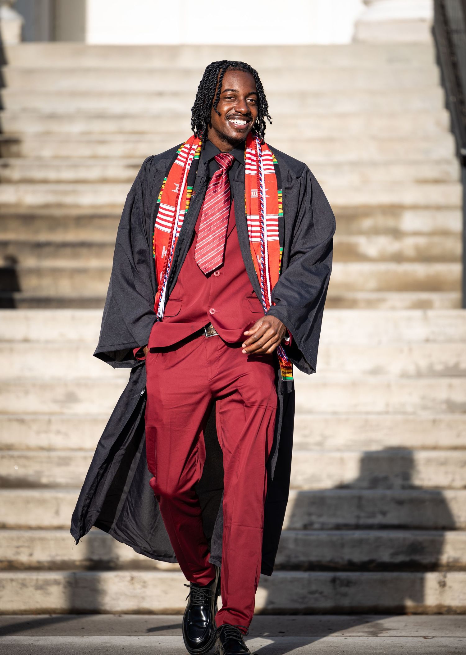 Graduate wearing burgundy suit and black graduation gown smiling on stone steps.