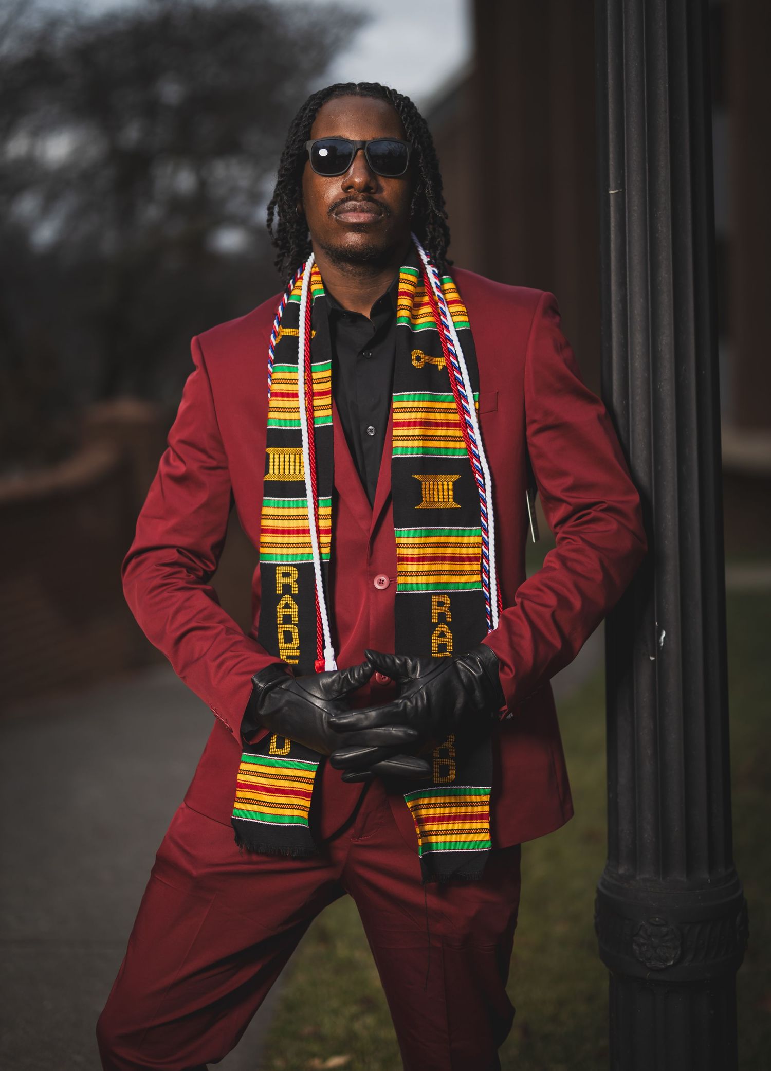 Professional in burgundy suit with kente scarf and camera standing by column.