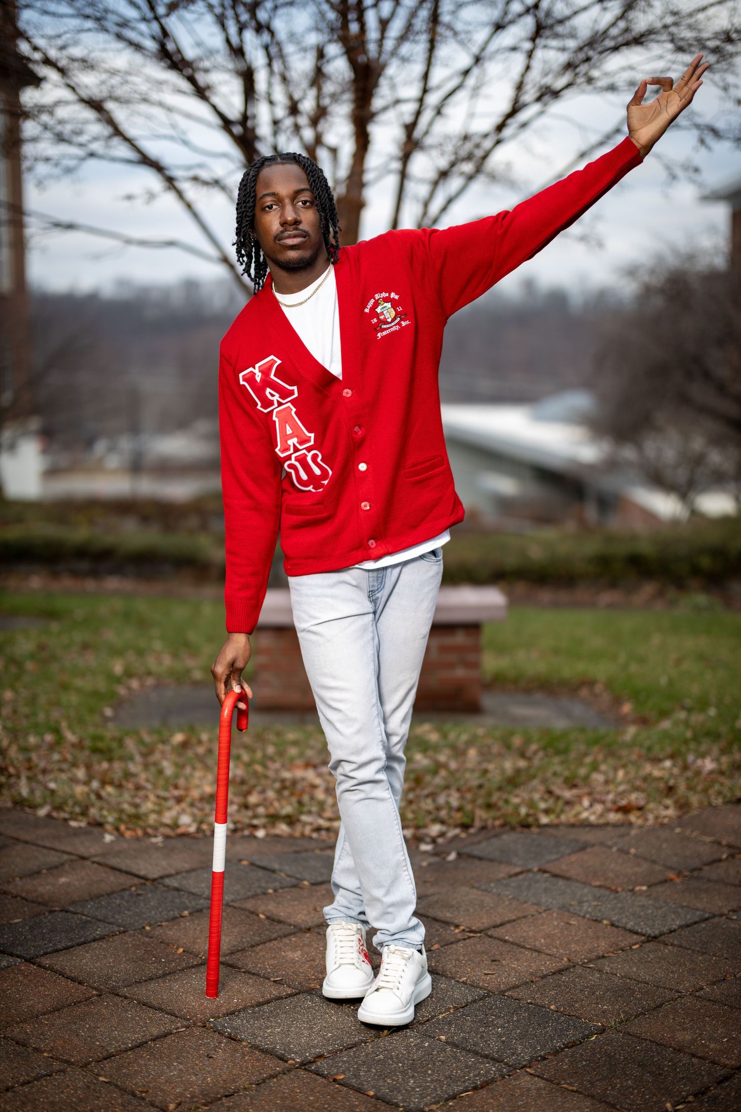 Graduate in red sweater and white pants striking pose with cane on brick pathway.