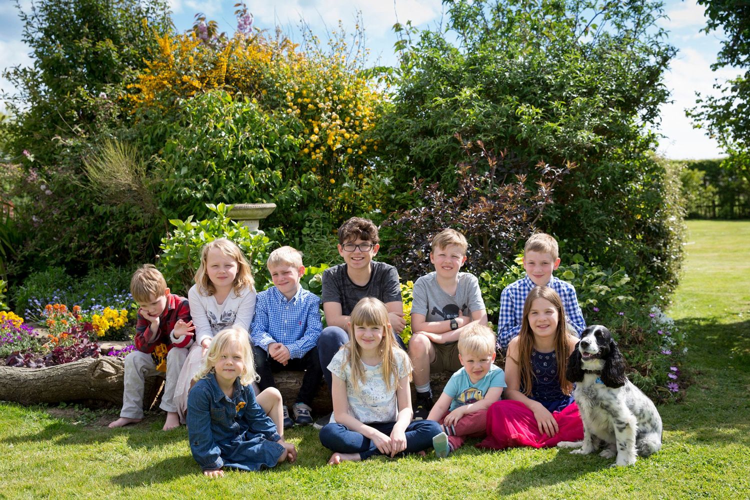 Large group sits on garden bench surrounded by blooming flowers and greenery.