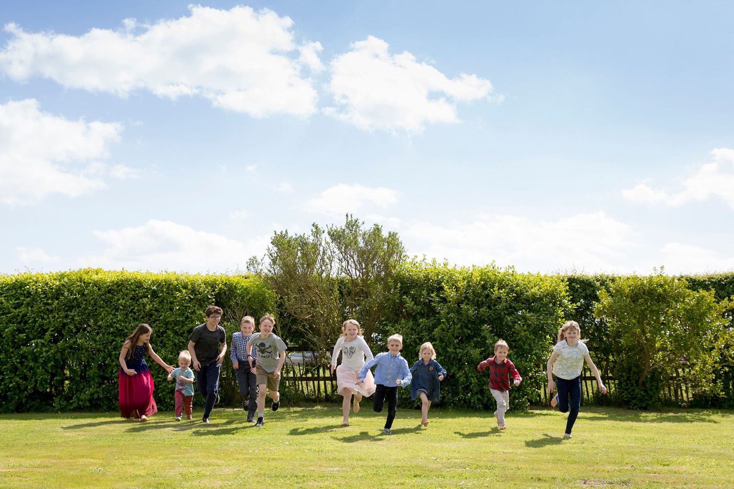 Children running and jumping on a grassy lawn with hedges and blue sky in the background.