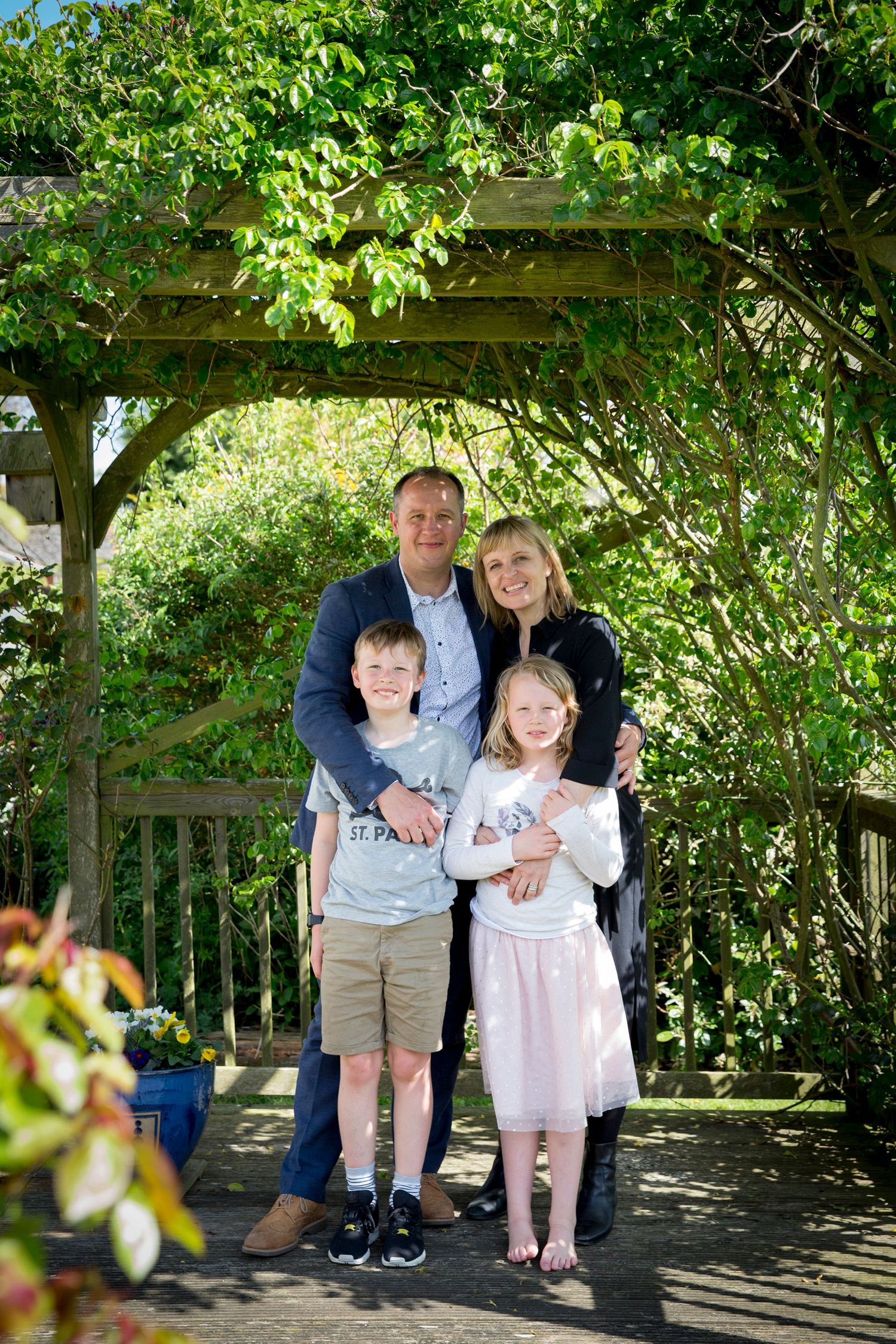 Family of four posing under a wooden garden pergola with green foliage.