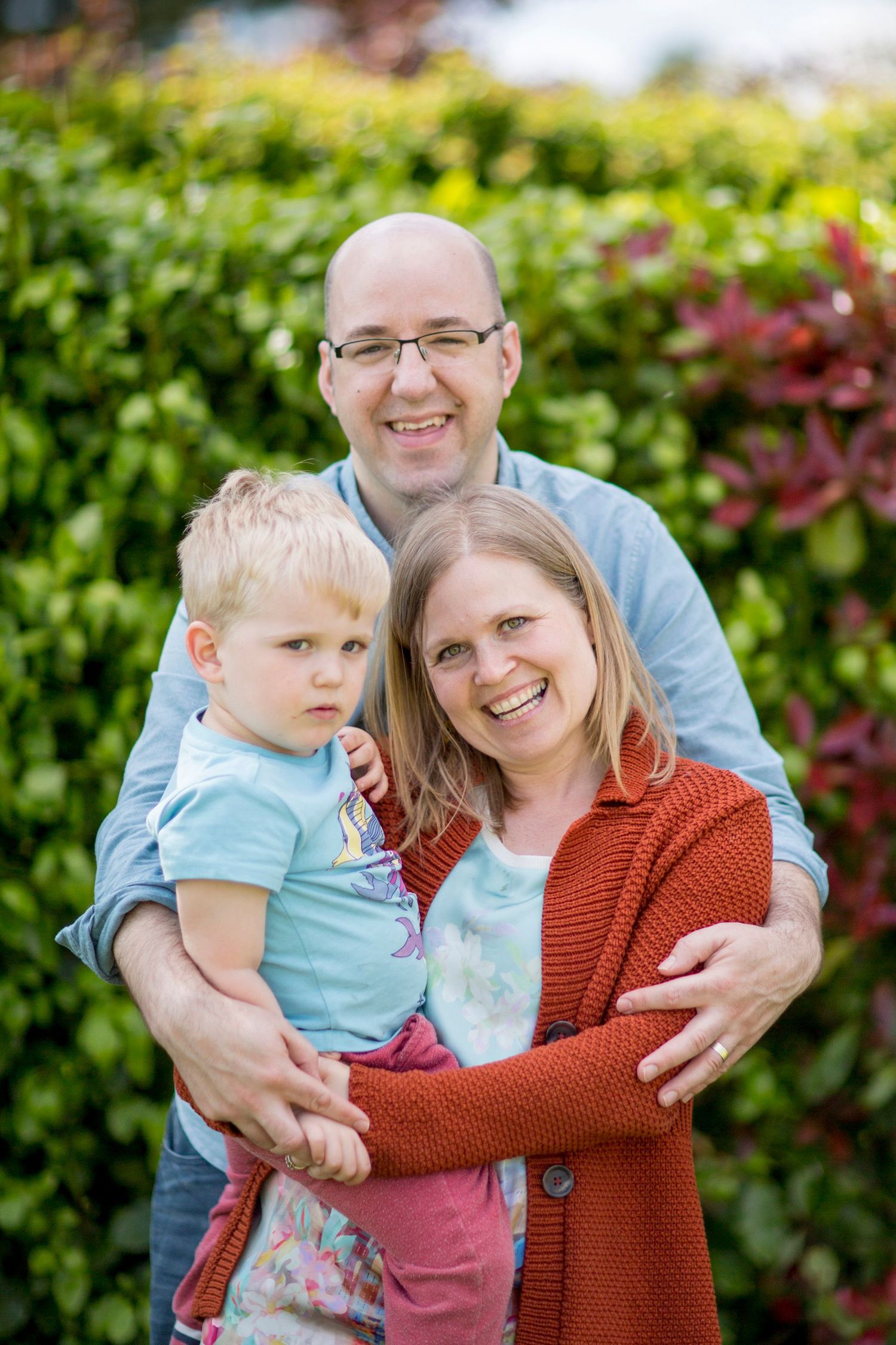 A family poses together outdoors in front of lush greenery on a sunny day.