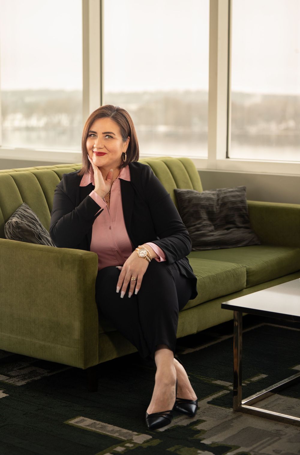 Professional in business attire seated on green velvet couch in modern office setting with large windows. Quad Cities Business Headshot Phot