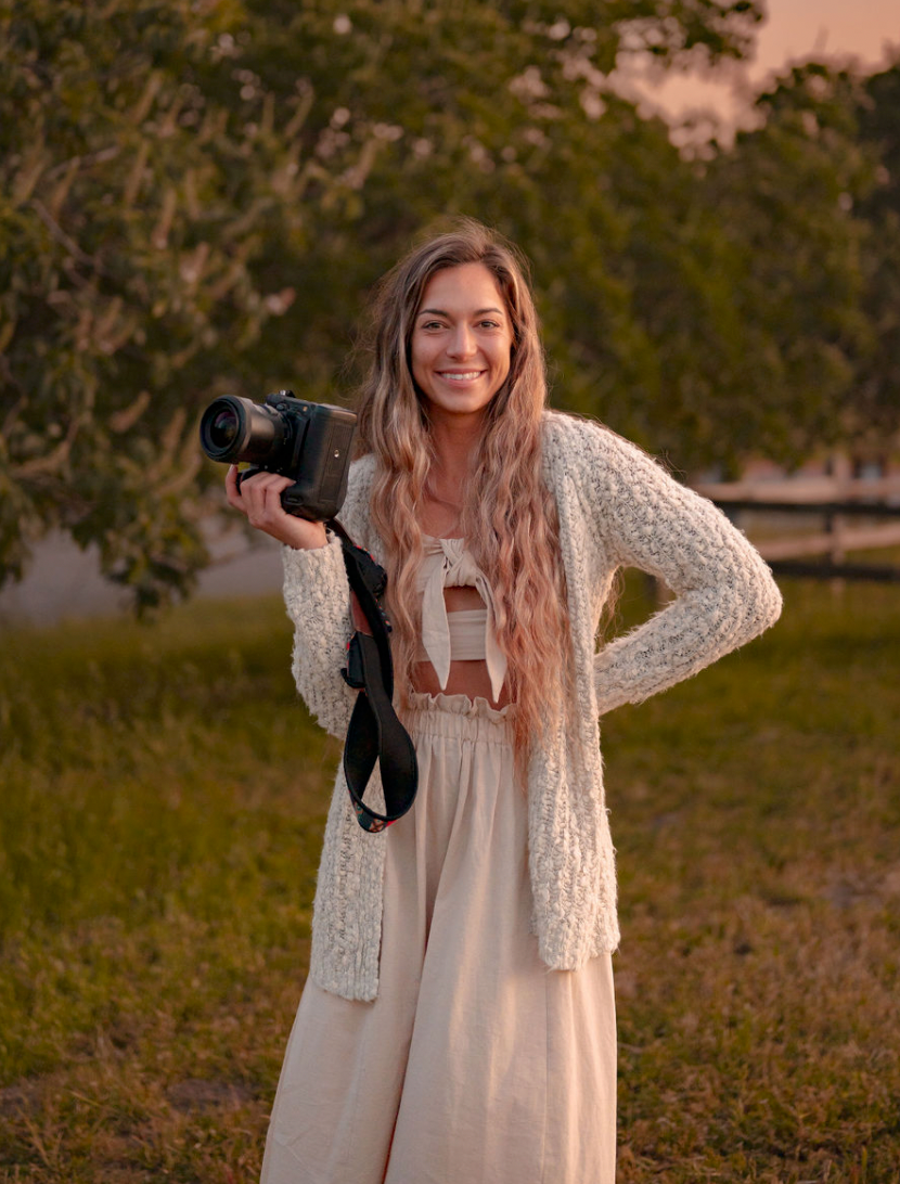 A photographer in a cream dress and knit cardigan holds a professional camera at golden hour.