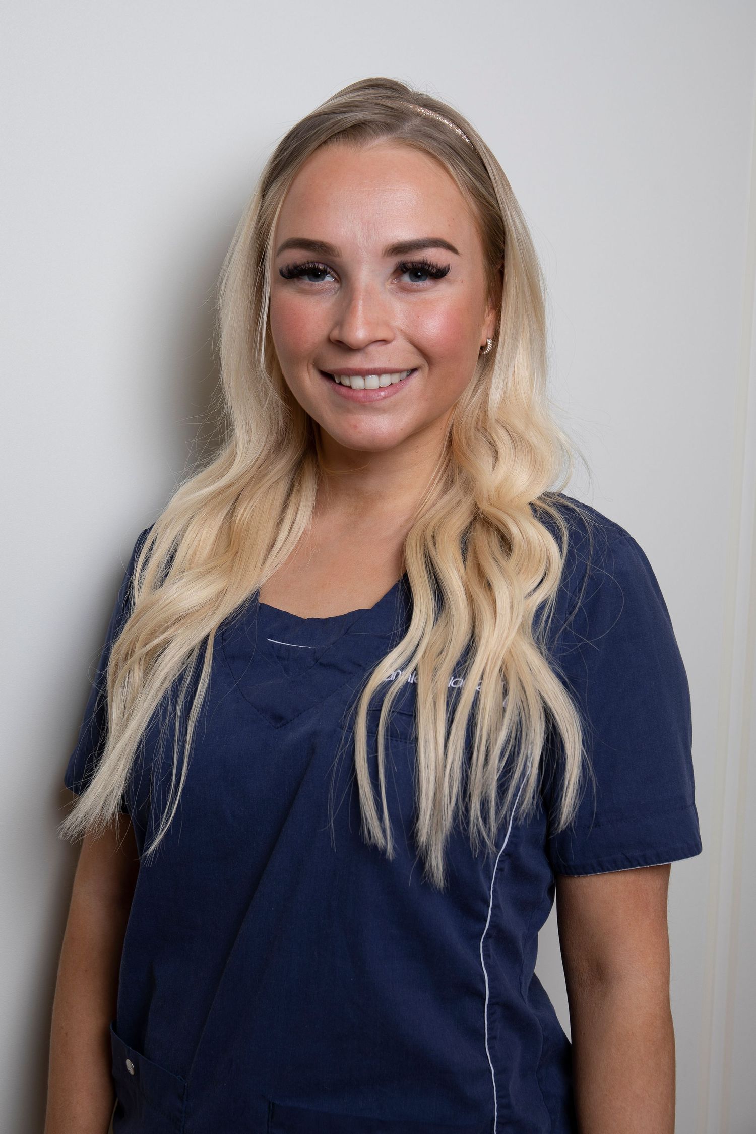 Young blonde woman smiling in navy blue medical scrubs with white trim against a white background.