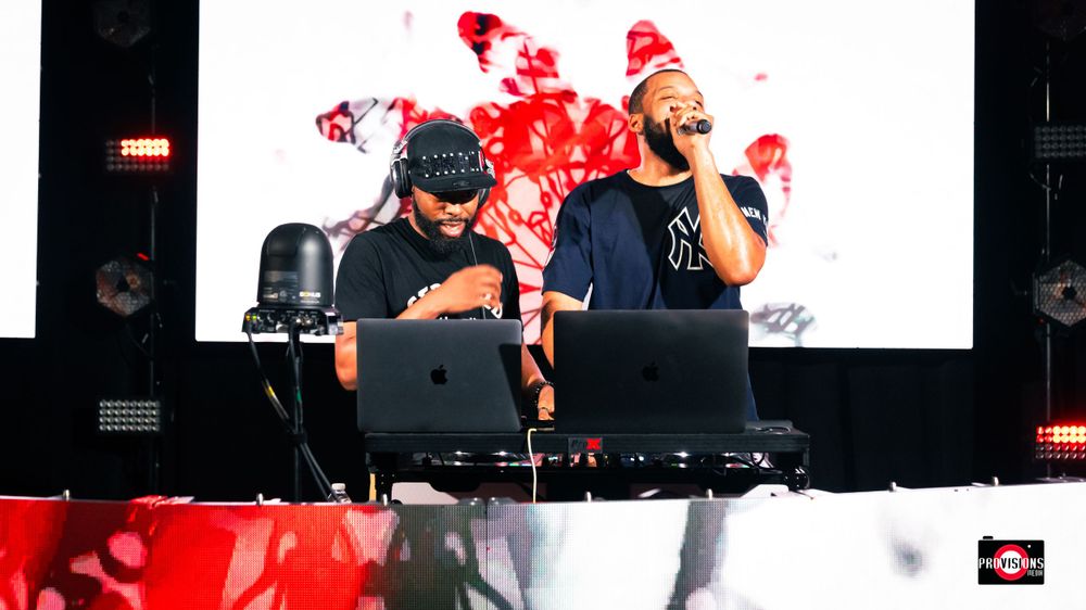 Two men performing as DJs on stage with laptops in front of a red and white backdrop at a live event.