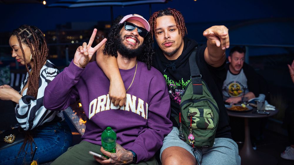 Two young men in streetwear pose together at a rooftop social event at night.