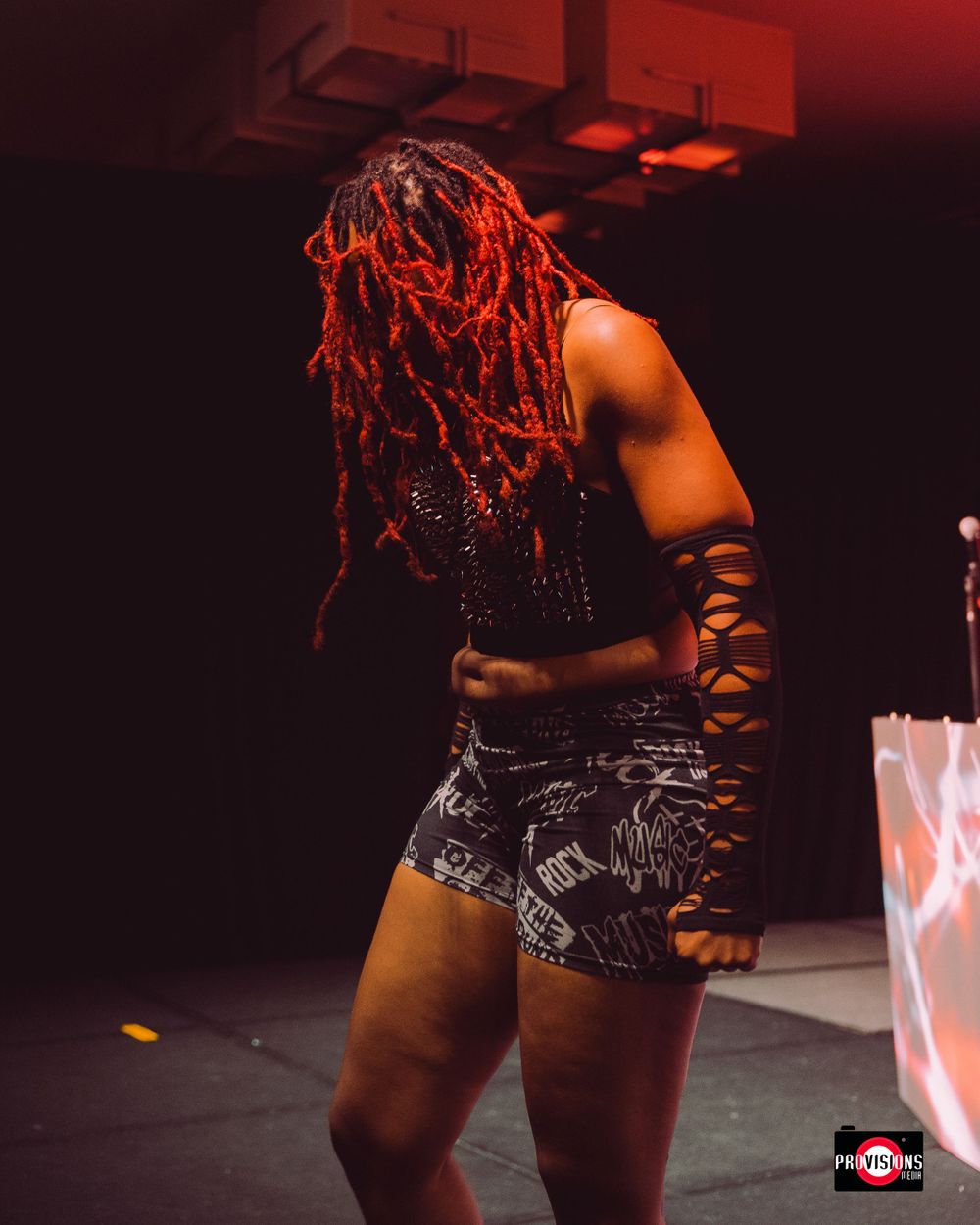 Performer with vibrant red dreadlocks wearing a black outfit dancing on a dimly lit stage.