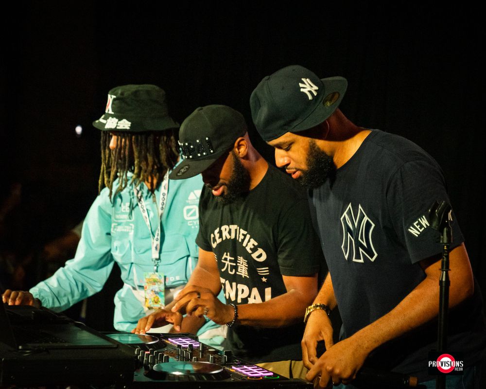 Three men working DJ equipment on a dark stage wearing caps and casual streetwear clothing.