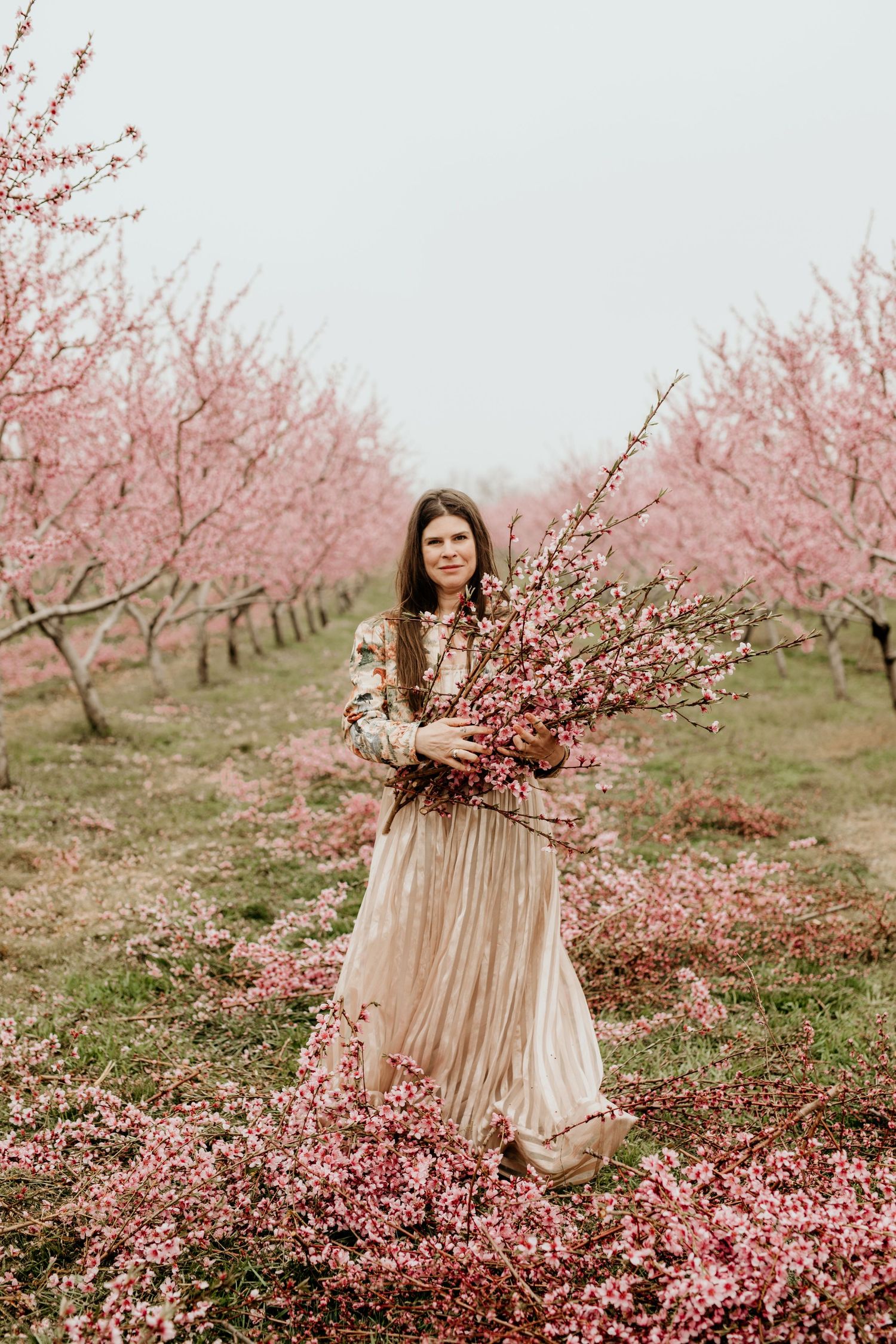 Woman in gold dress holding pink cherry blossom branches in blooming orchard.