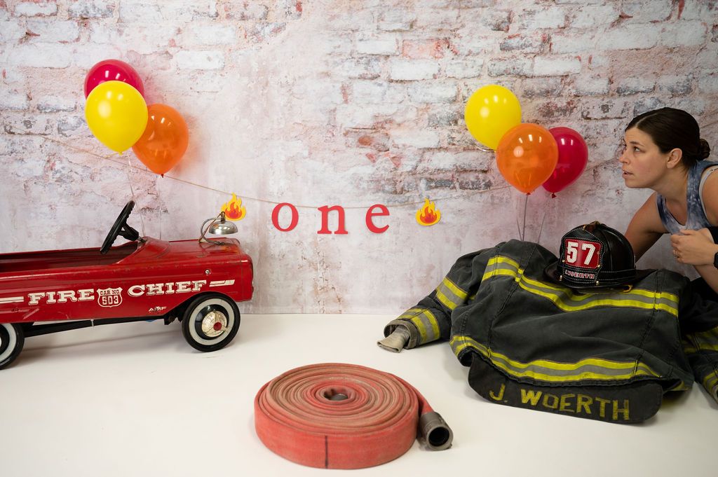 First birthday setup with red wagon, fire chief theme, and colorful balloons against brick wall.