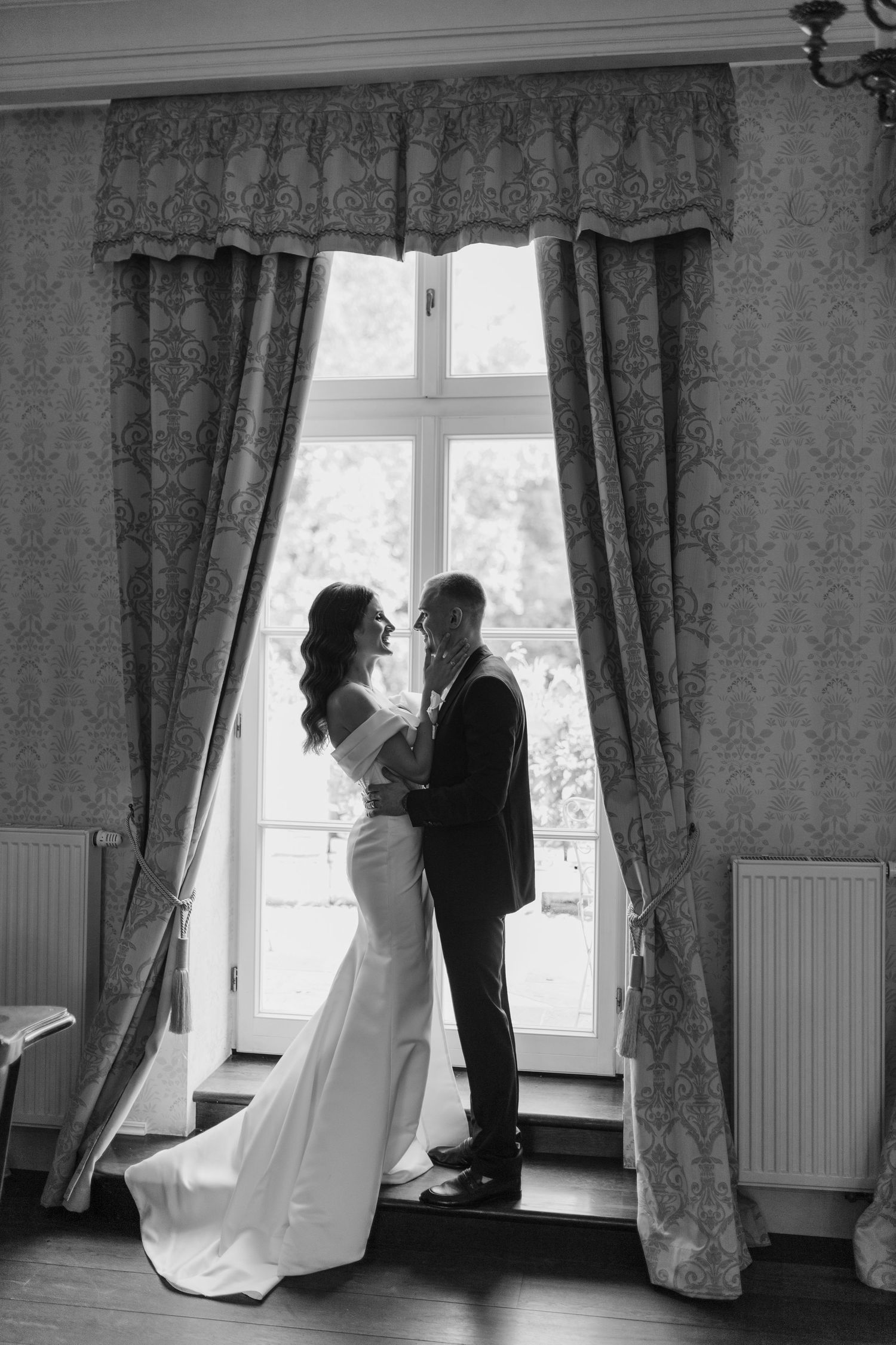 Romantic black and white photo of couple by window with elegant curtains.