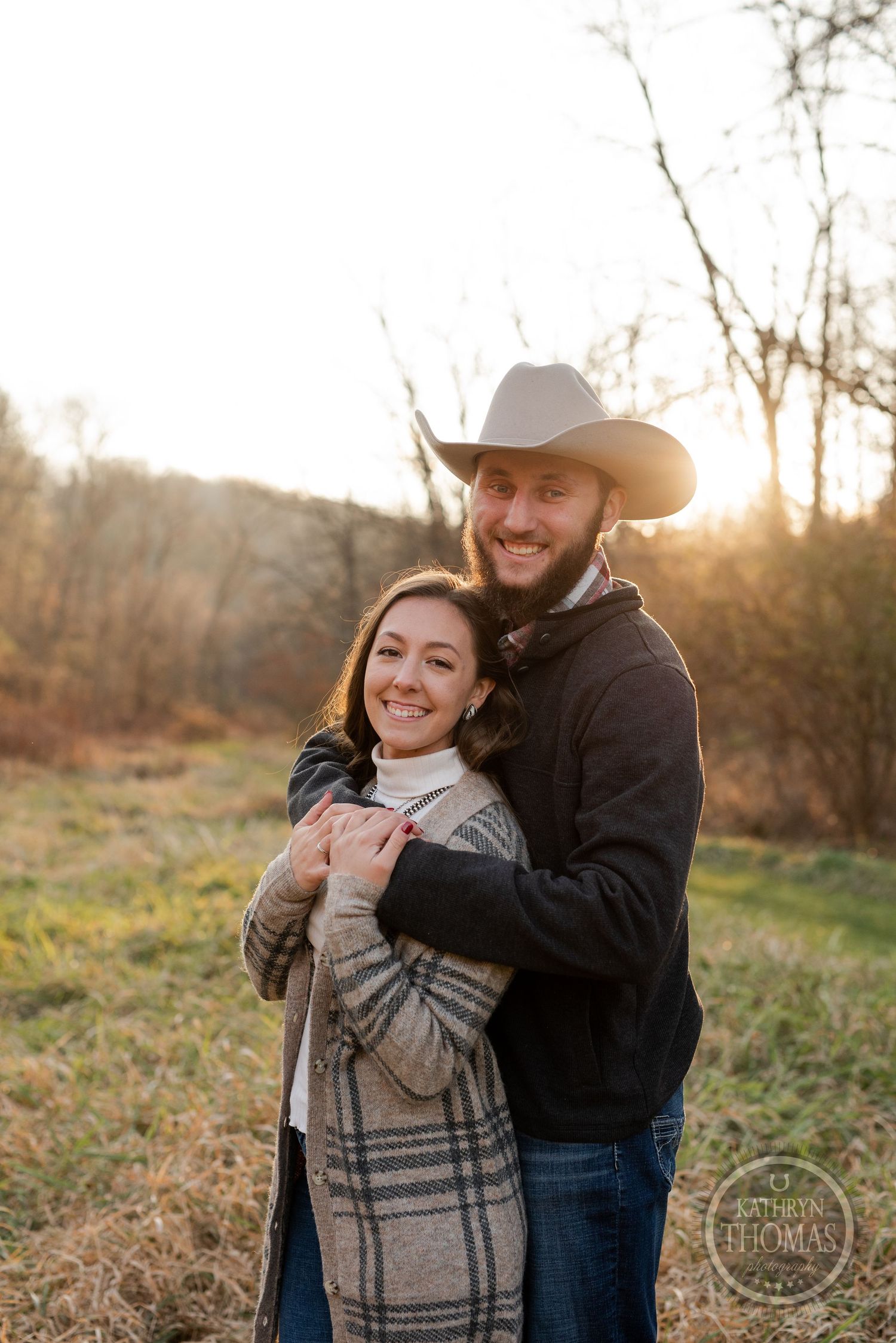 A happy couple embraces in a field at sunset wearing casual western attire.