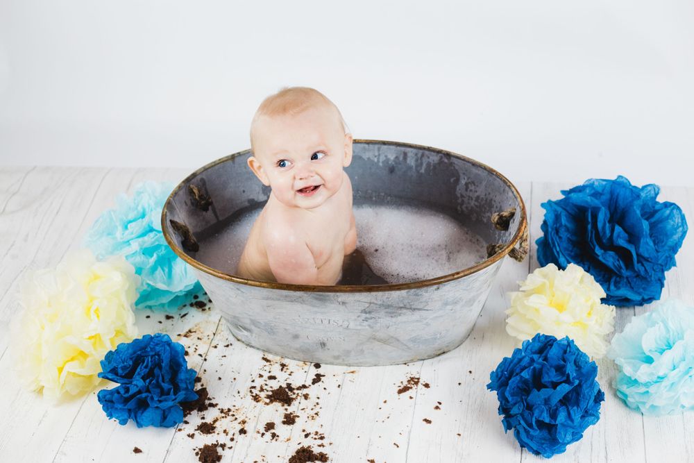Baby sitting in vintage metal tub surrounded by blue and teal paper flowers during bath photoshoot.