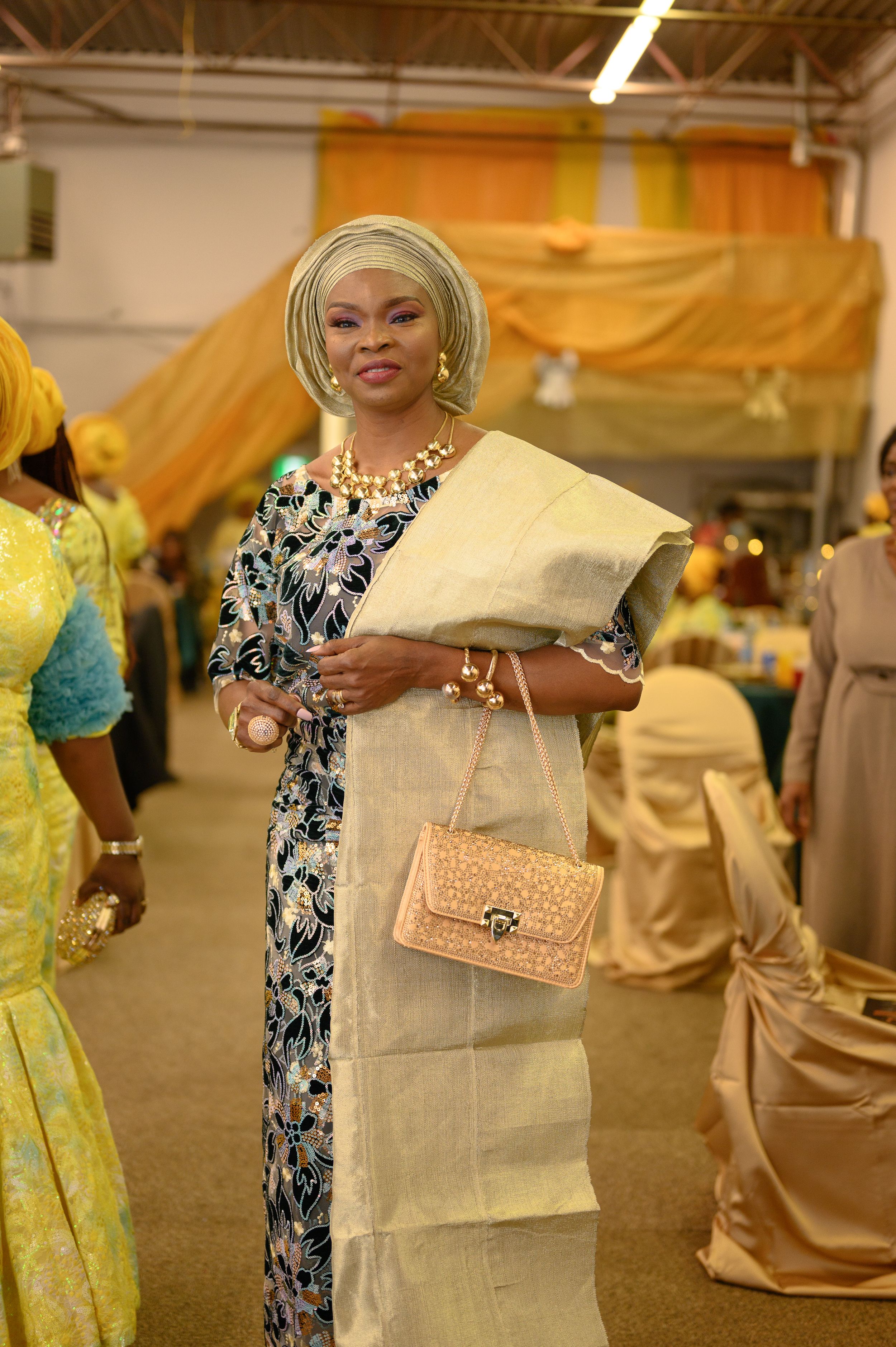Elegant guest in traditional Nigerian dress and gele holding a gift at formal event.
