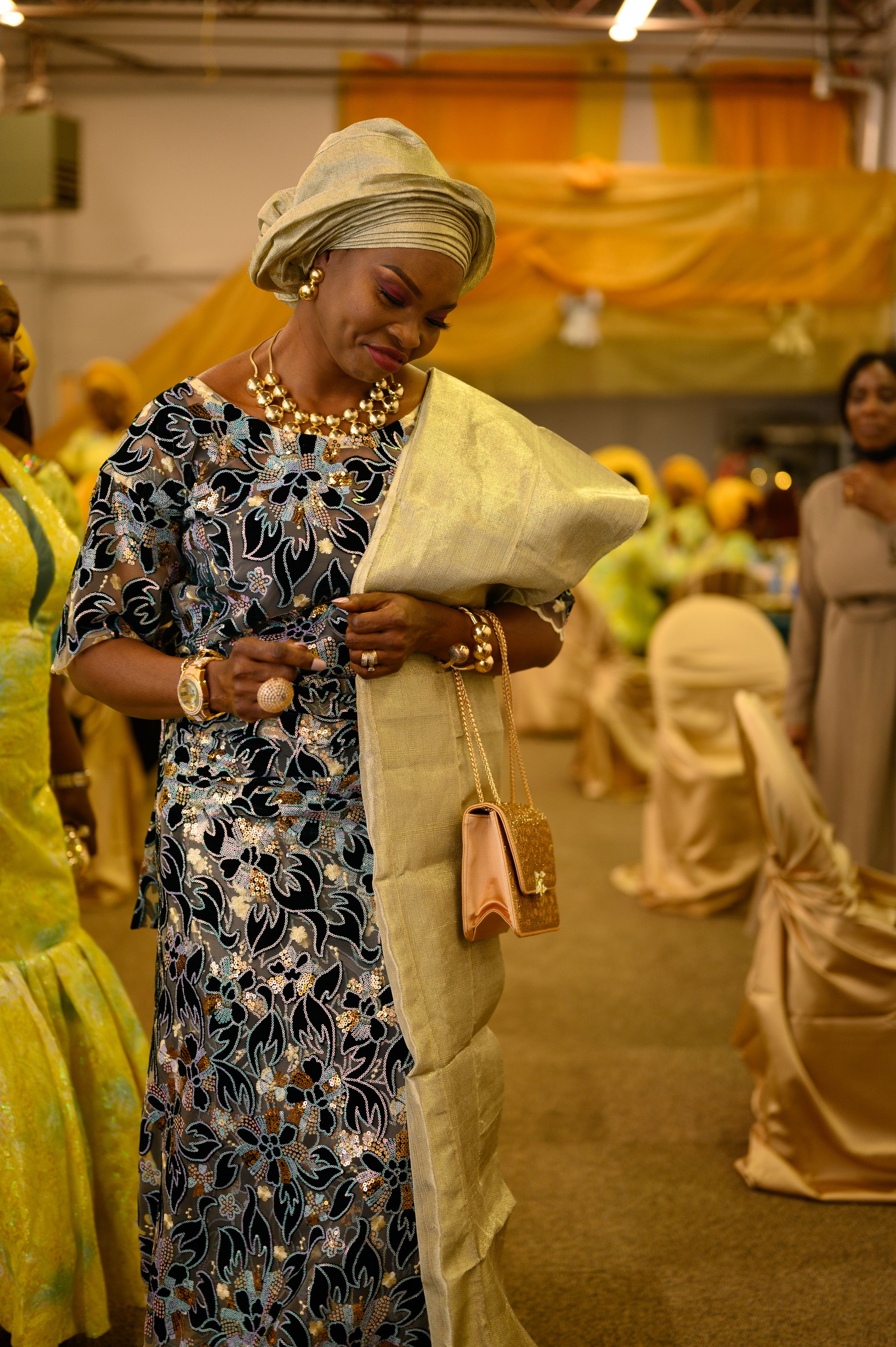 Guest in blue floral dress and gele headwrap smiling while holding gift bag at celebration.