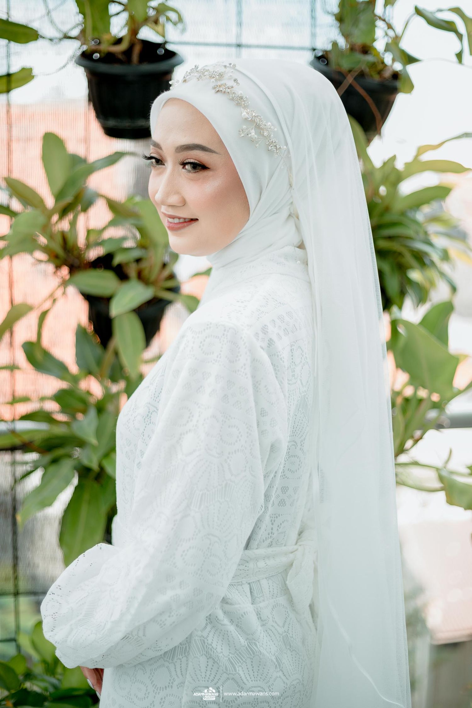 A bride in a white hijab and embellished dress poses among green plants in a sunlit setting.