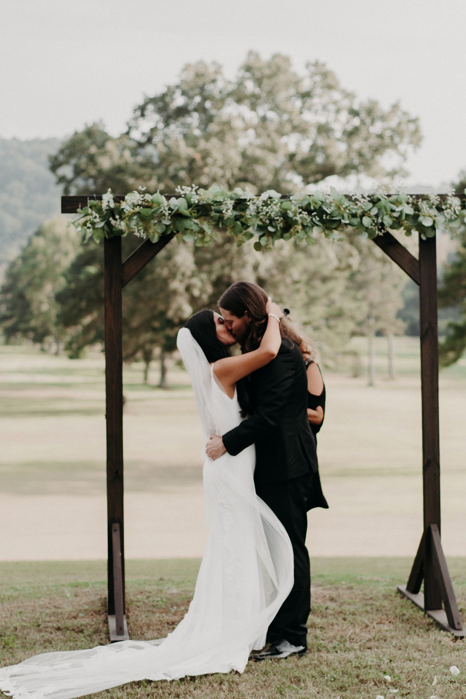 Newlyweds share an intimate moment under a wooden arch decorated with white flowers and greenery in an outdoor setting.