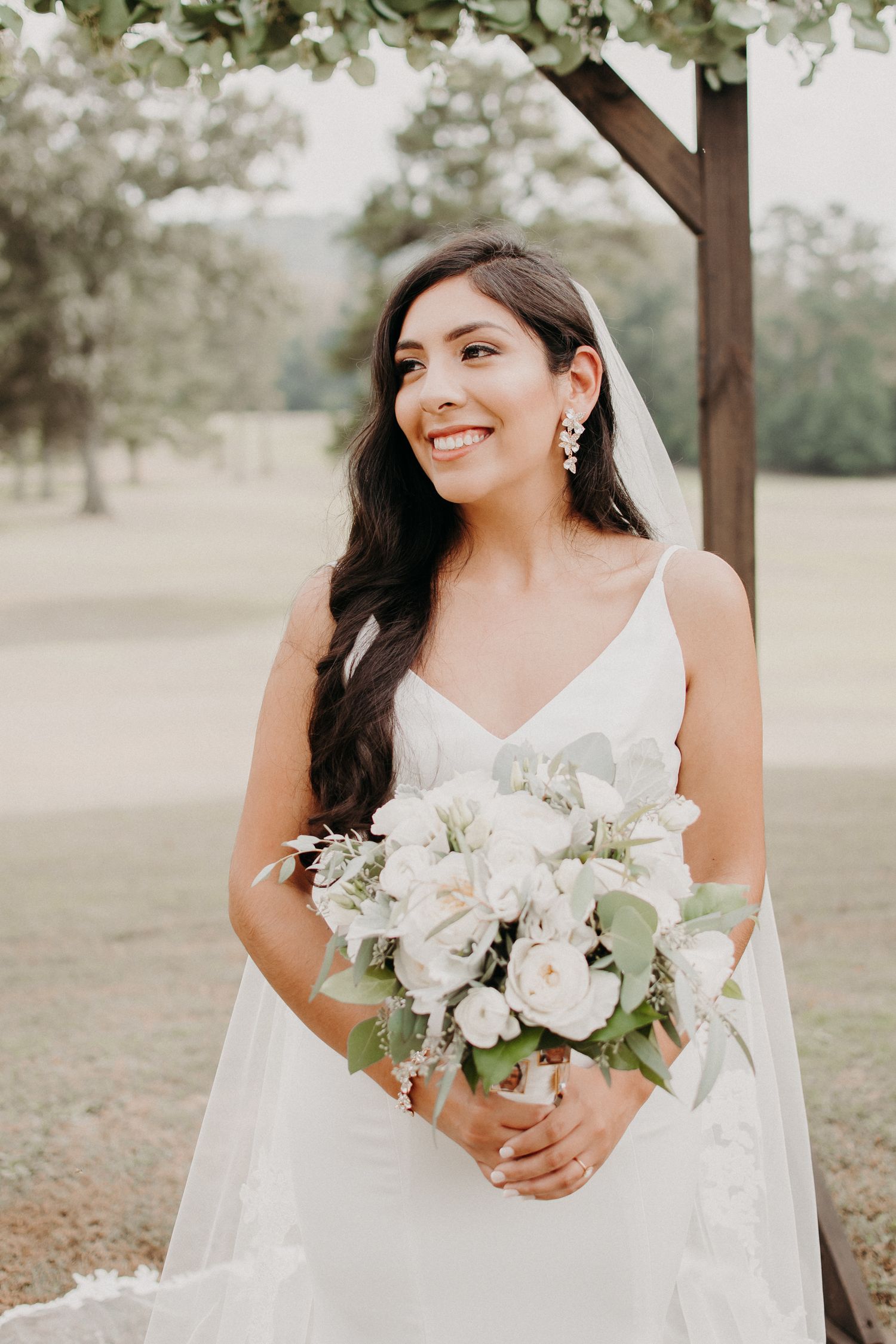 Elegant bride holding white bouquet under rustic wooden arch decorated with flowers.