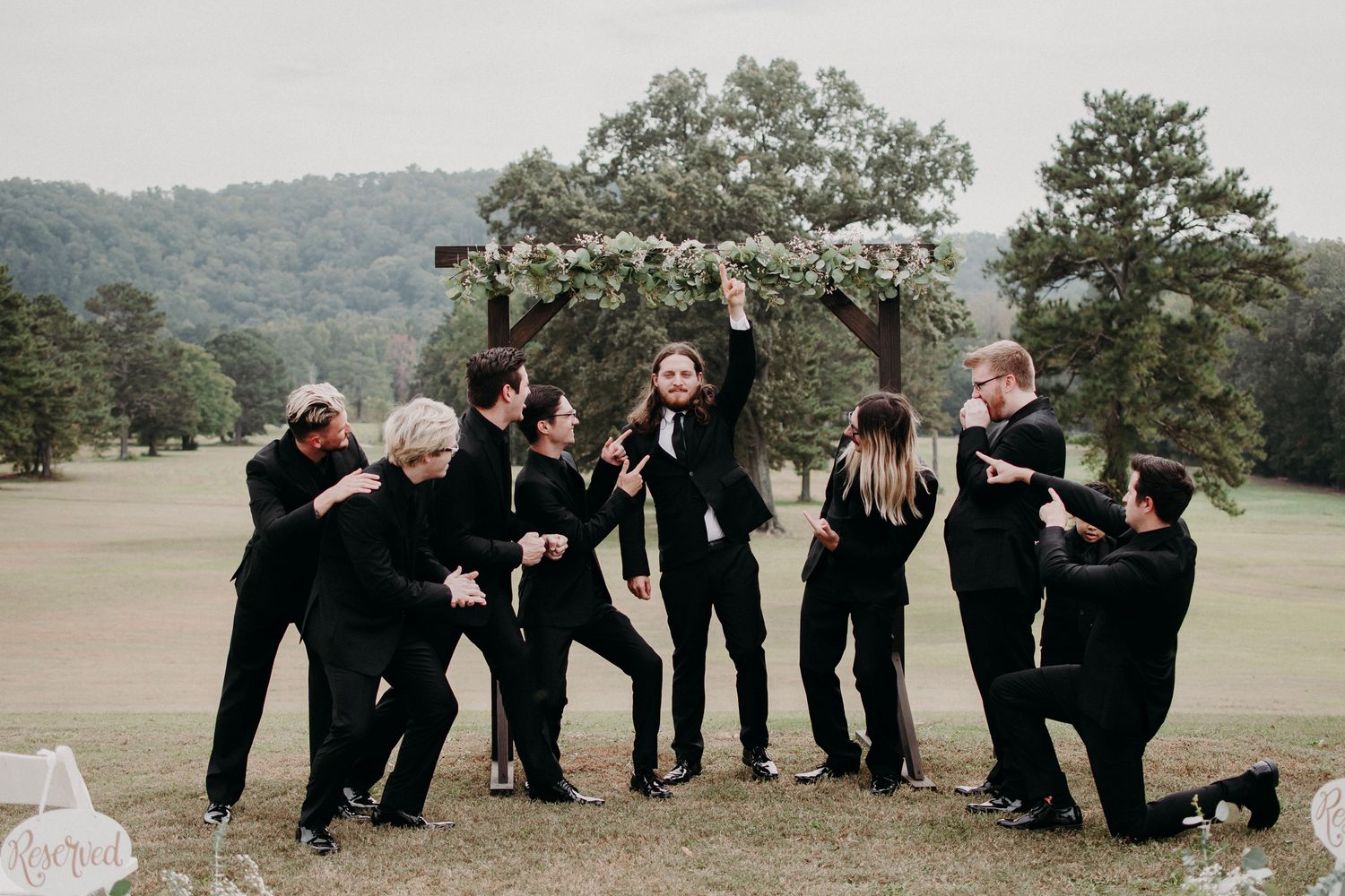 Wedding party members in black attire strike fun poses under floral arch.