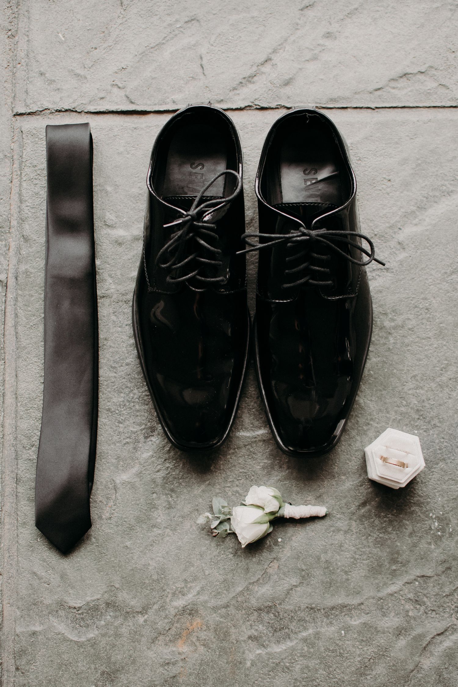 Groom's formal accessories including black dress shoes, tie, and boutonniere on gray surface.