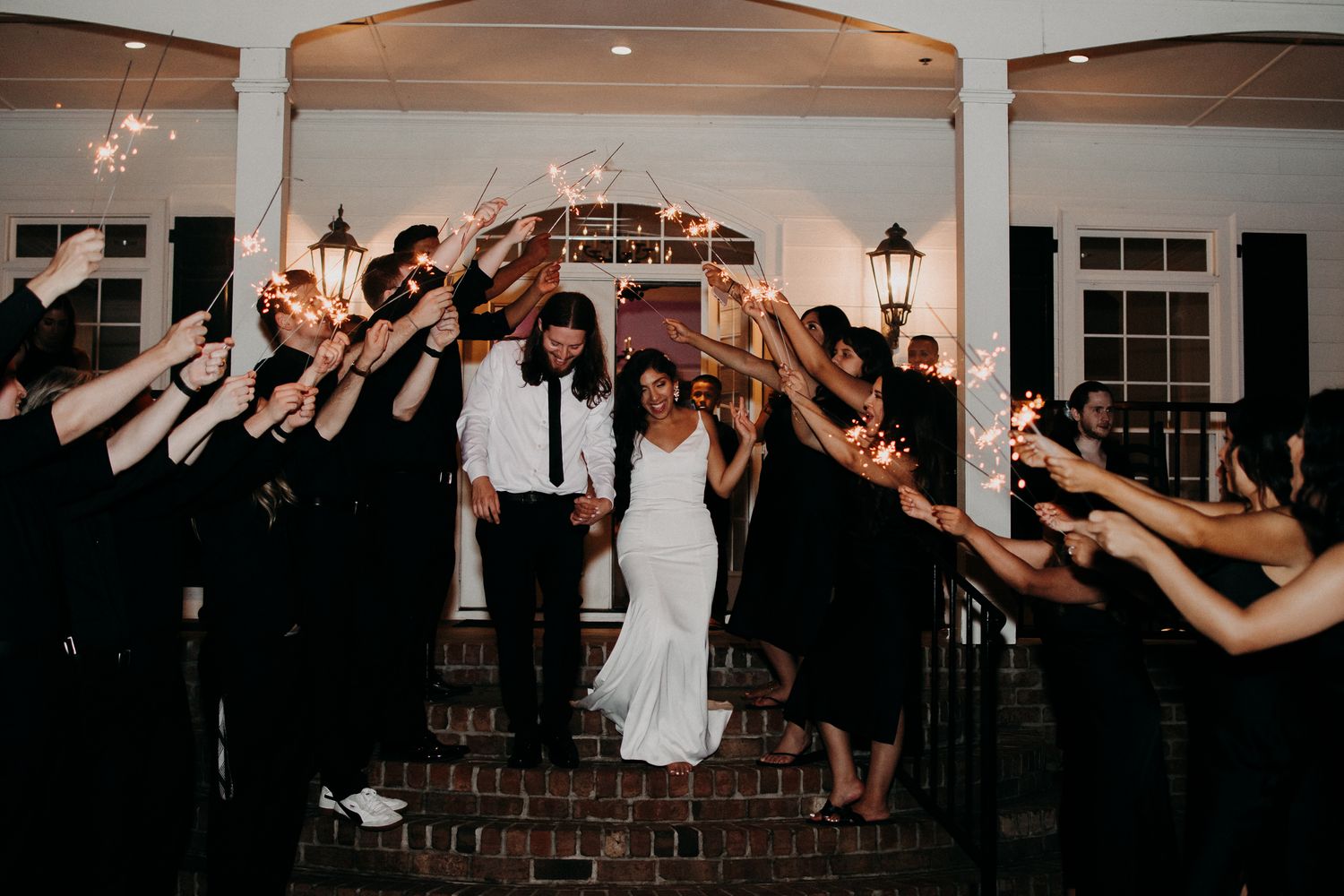 Wedding guests form a sparkler tunnel exit for the newlyweds on brick steps.