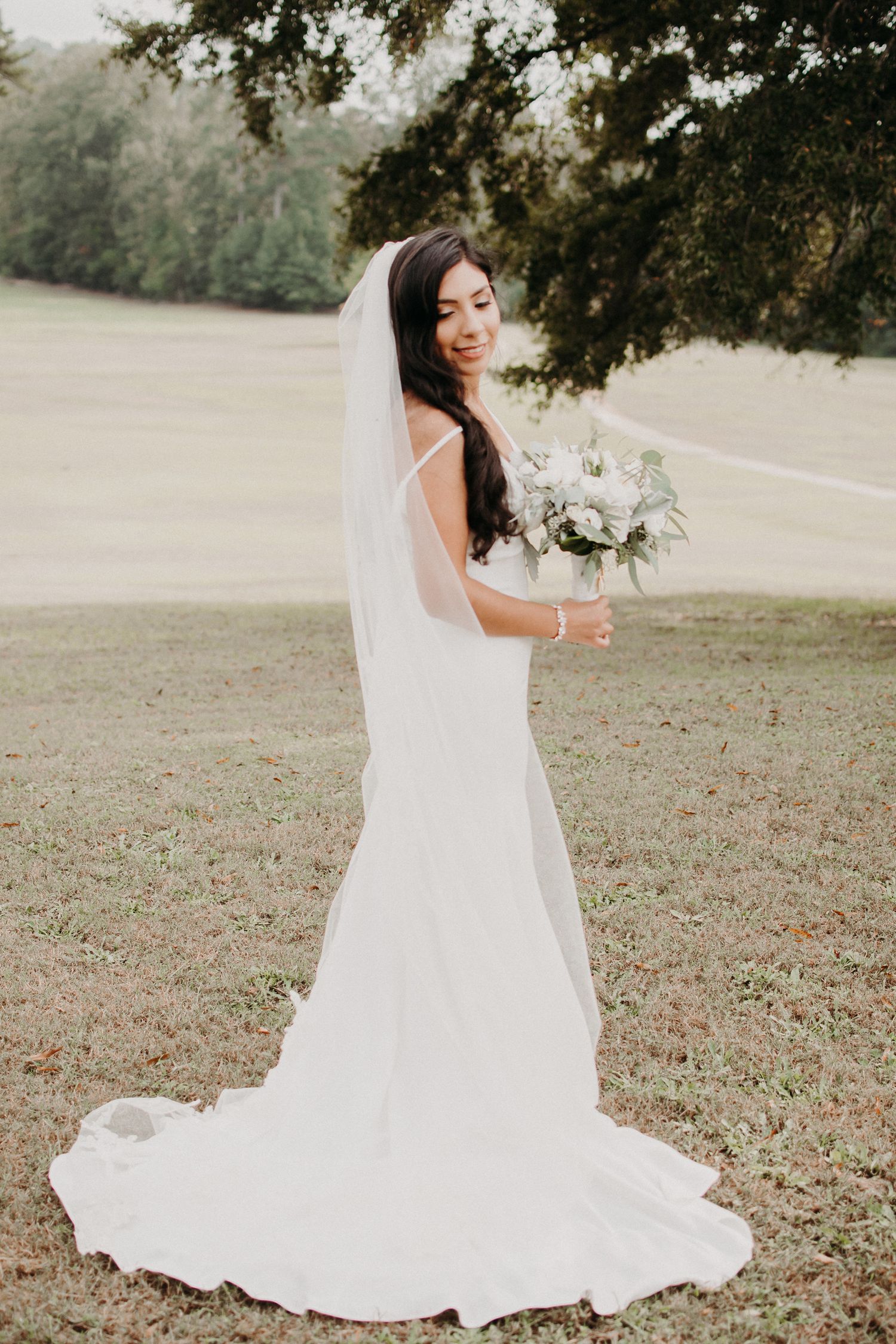 Full-length bridal portrait in sleek white gown with long train and veil in outdoor setting.