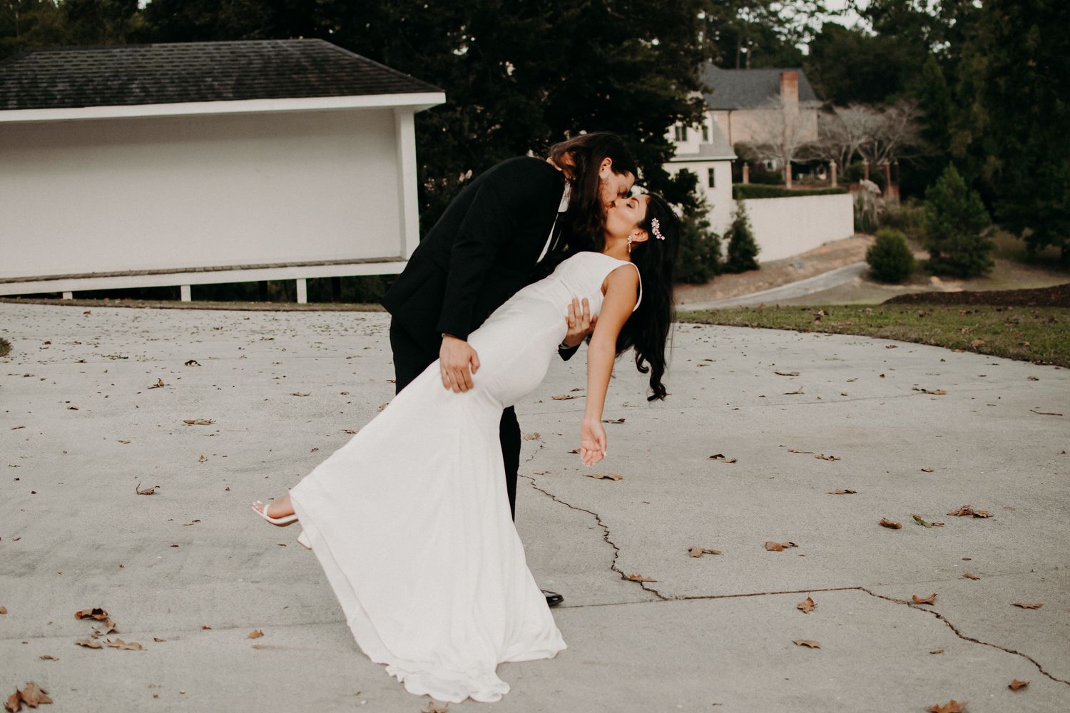 A couple performs a romantic dip pose in formal wedding attire on a driveway with fallen leaves.