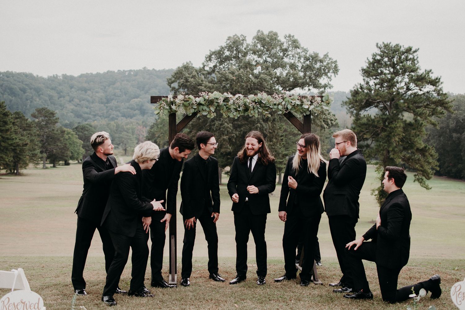 Wedding party in black attire poses playfully under floral arch in outdoor setting.