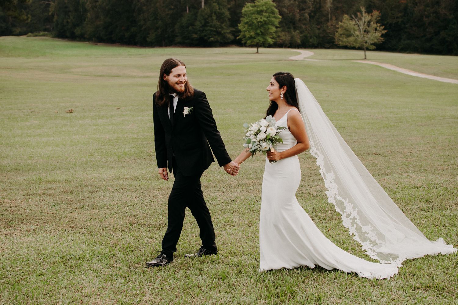 Couple walking hand in hand across grassy field during wedding photoshoot.