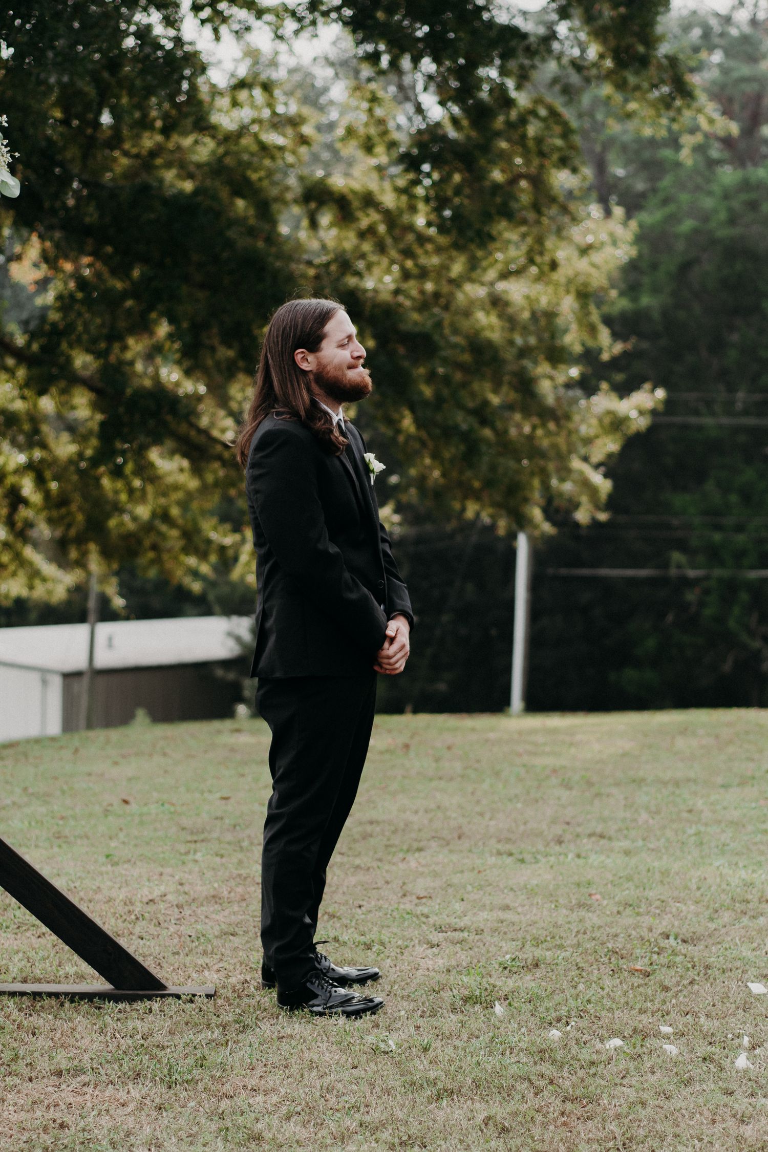 A person in a black suit stands thoughtfully in a grassy outdoor setting.