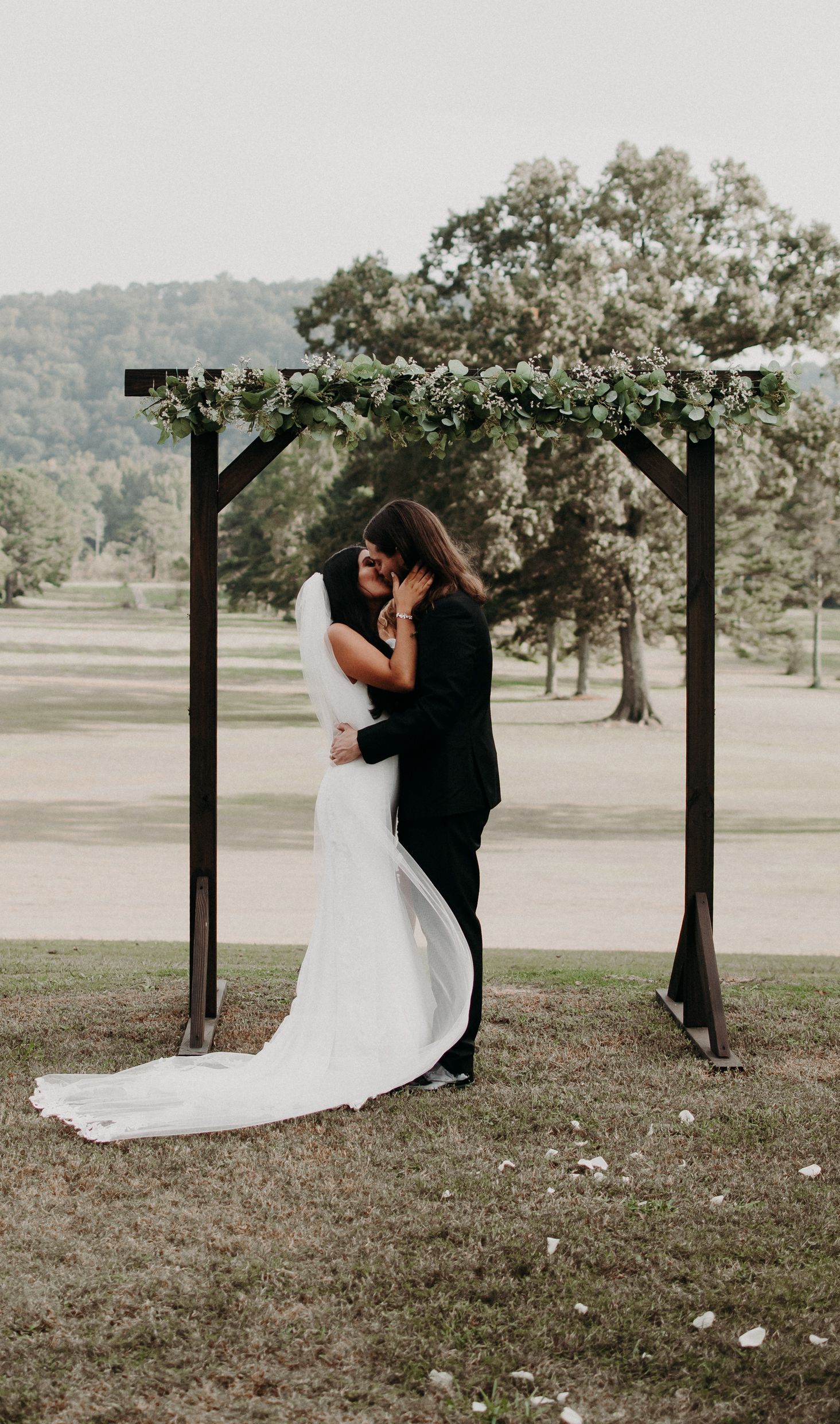 A romantic embrace under a floral-decorated wooden arch at an outdoor wedding.