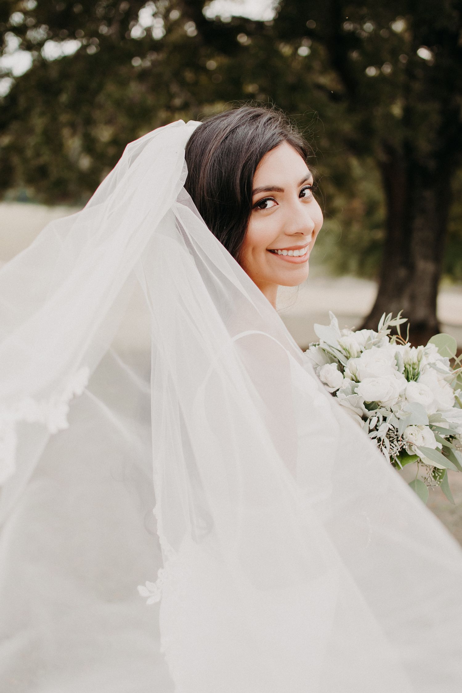 Side profile bridal portrait with flowing veil and white bouquet against natural backdrop.