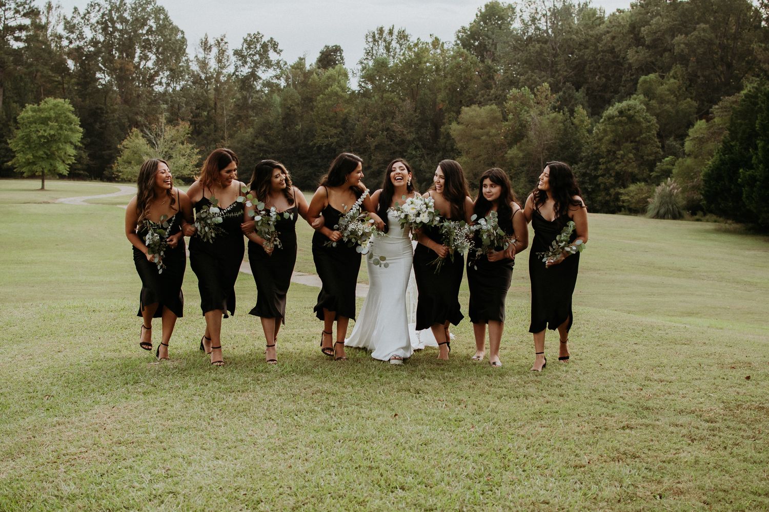 Wedding party of bridesmaids in black dresses laugh together on a grassy lawn.