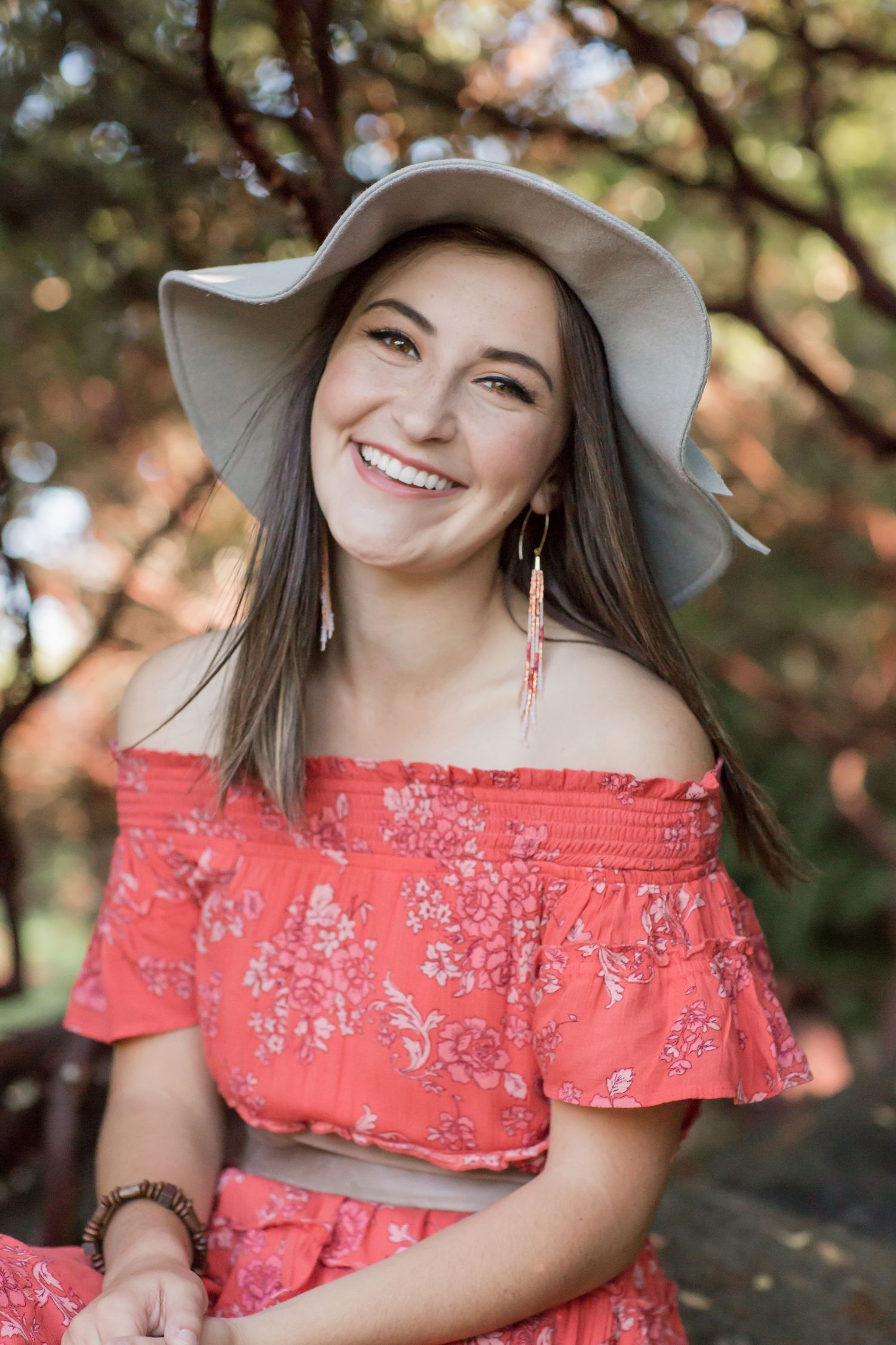 Cheerful woman in coral floral off-shoulder top wearing a wide-brim gray hat outdoors.