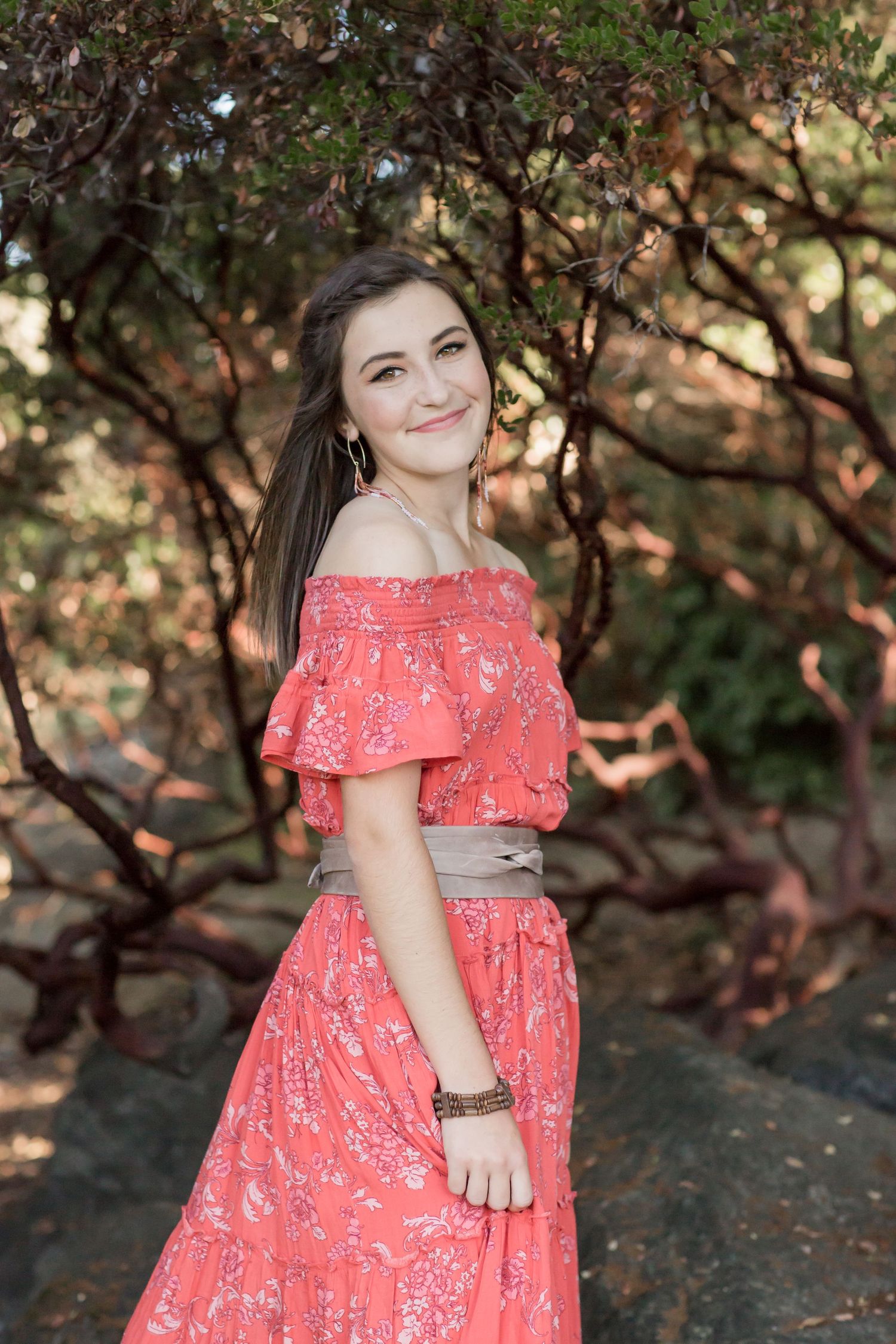 Brunette woman in coral off-shoulder floral maxi dress smiling among twisted trees outdoors.