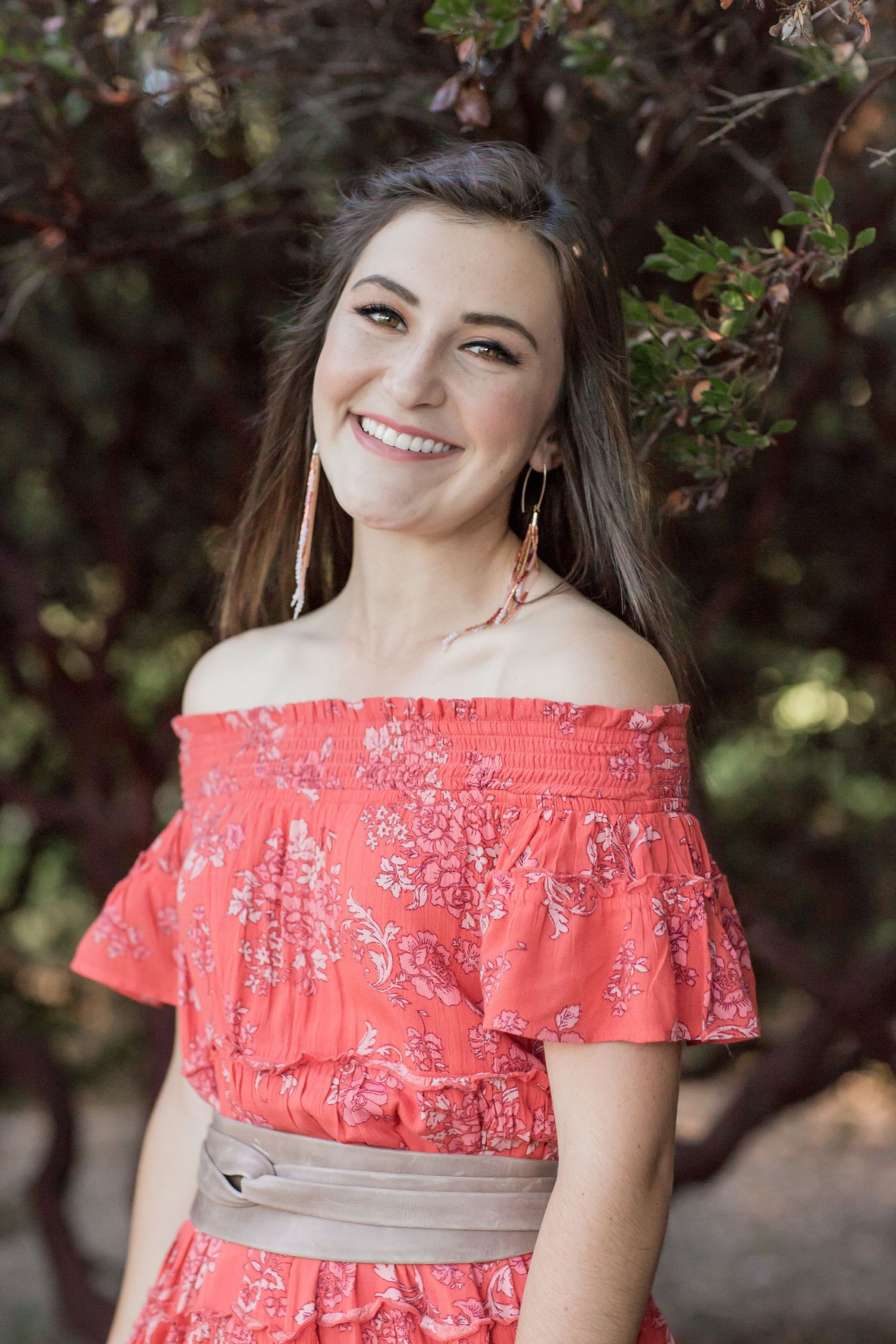 Smiling young woman in coral off-shoulder floral dress with gold earrings posing outdoors.