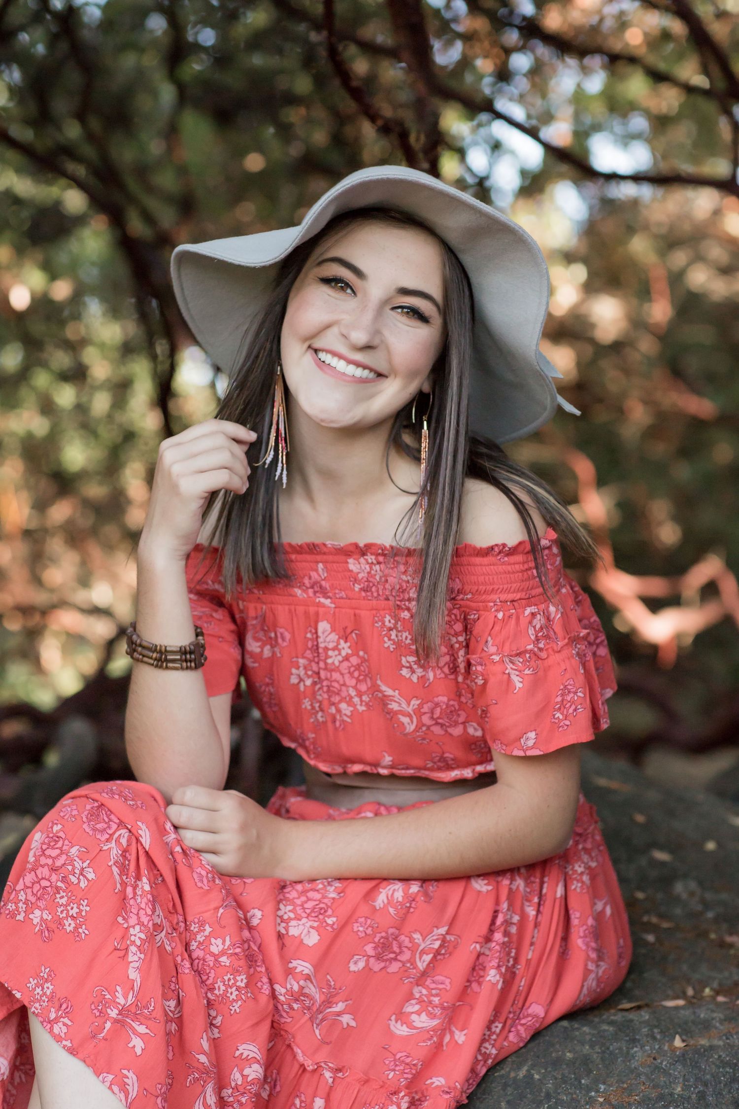 Young woman in coral floral off-shoulder dress and wide-brim hat sitting on rocks outdoors.