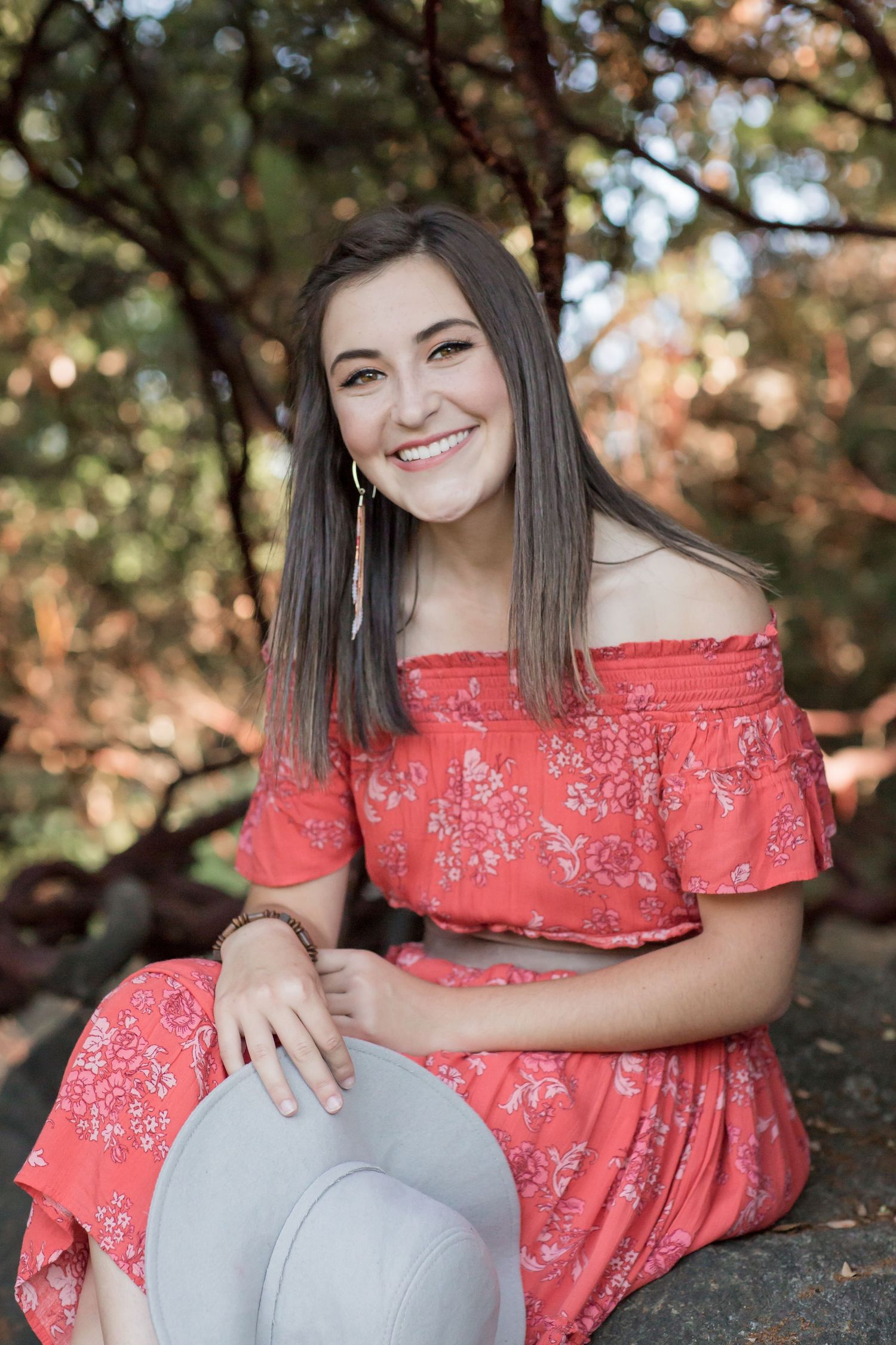 Smiling brunette woman in coral off-shoulder floral dress sitting outdoors holding a gray hat.