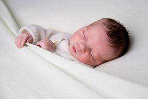Sleeping newborn in white onesie resting peacefully on light bedding.