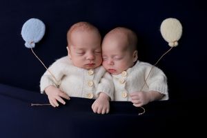 Twin newborns in white sweaters sleeping peacefully with small balloons against a dark background.